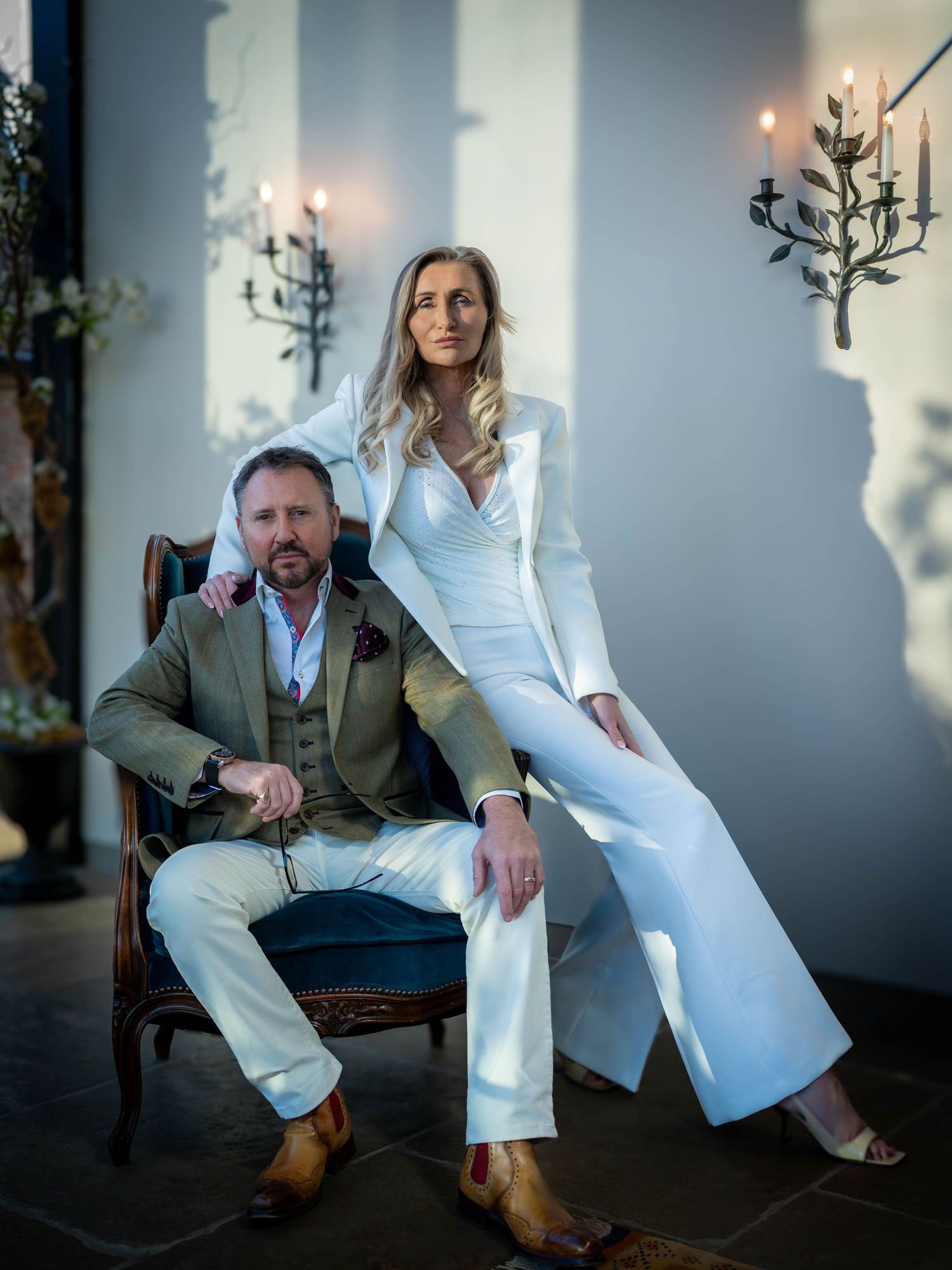 Elegant couple posing in a candlelit room, man in olive suit seated, woman in white jumpsuit standing beside him.