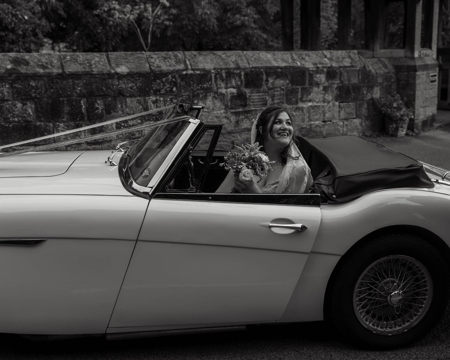 Bride smiling in a classic white Austin-Healey convertible sports car, black and white wedding photography.