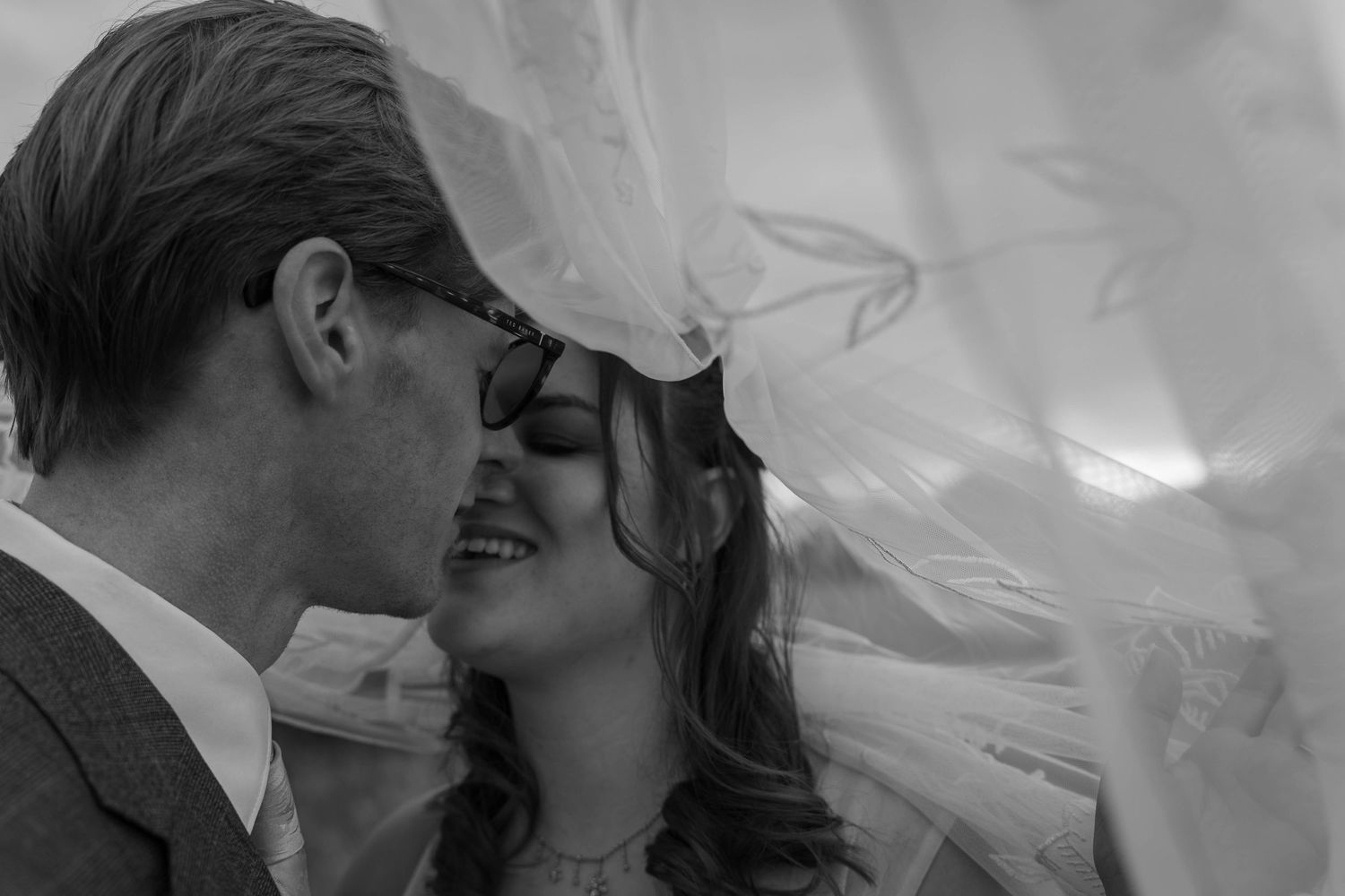 Black and white photo of a bride and groom sharing a romantic moment under a flowing wedding veil.