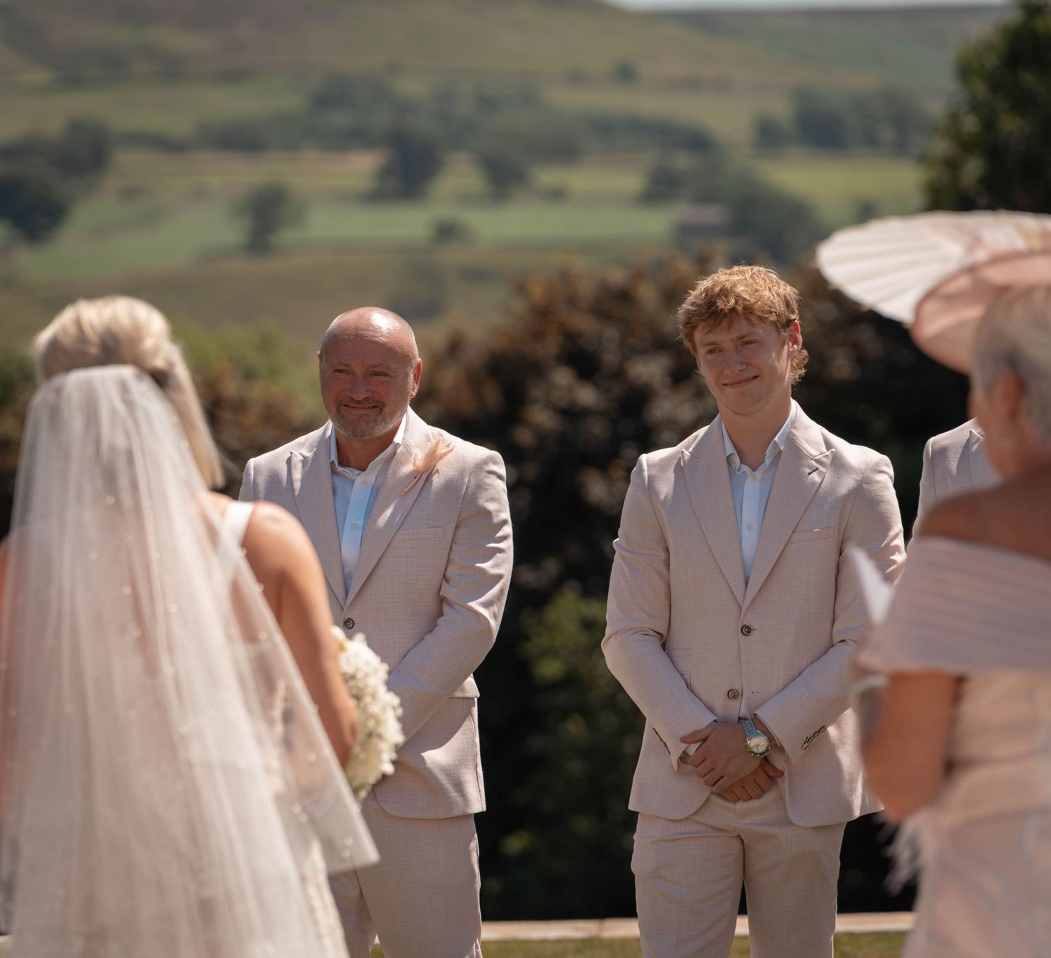 Groom smiling as bride walks down the aisle at an outdoor wedding ceremony with rolling green hills in the background.