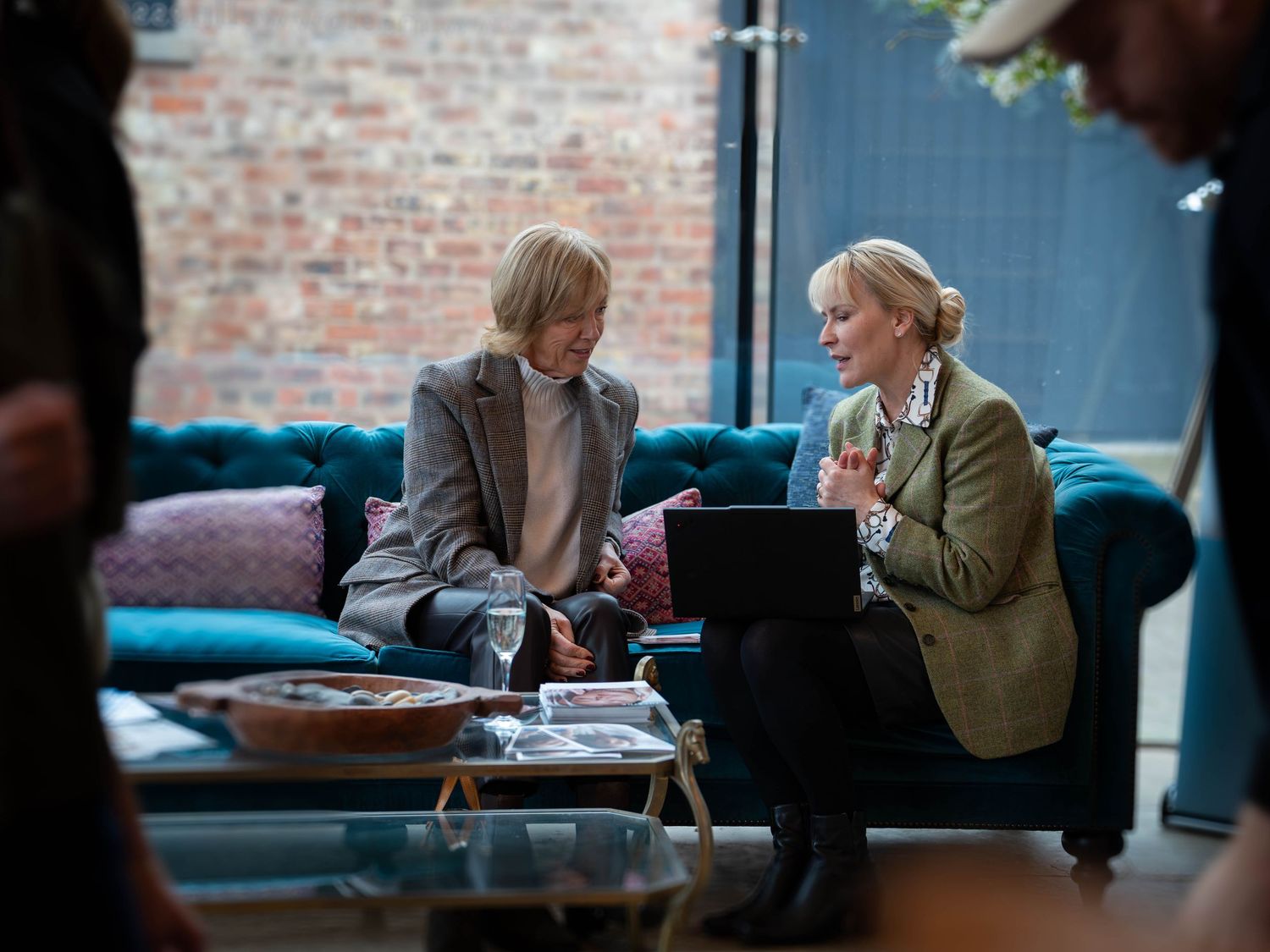 Two women in blazers discuss work on a laptop while seated on a teal sofa in a modern brick-walled office.