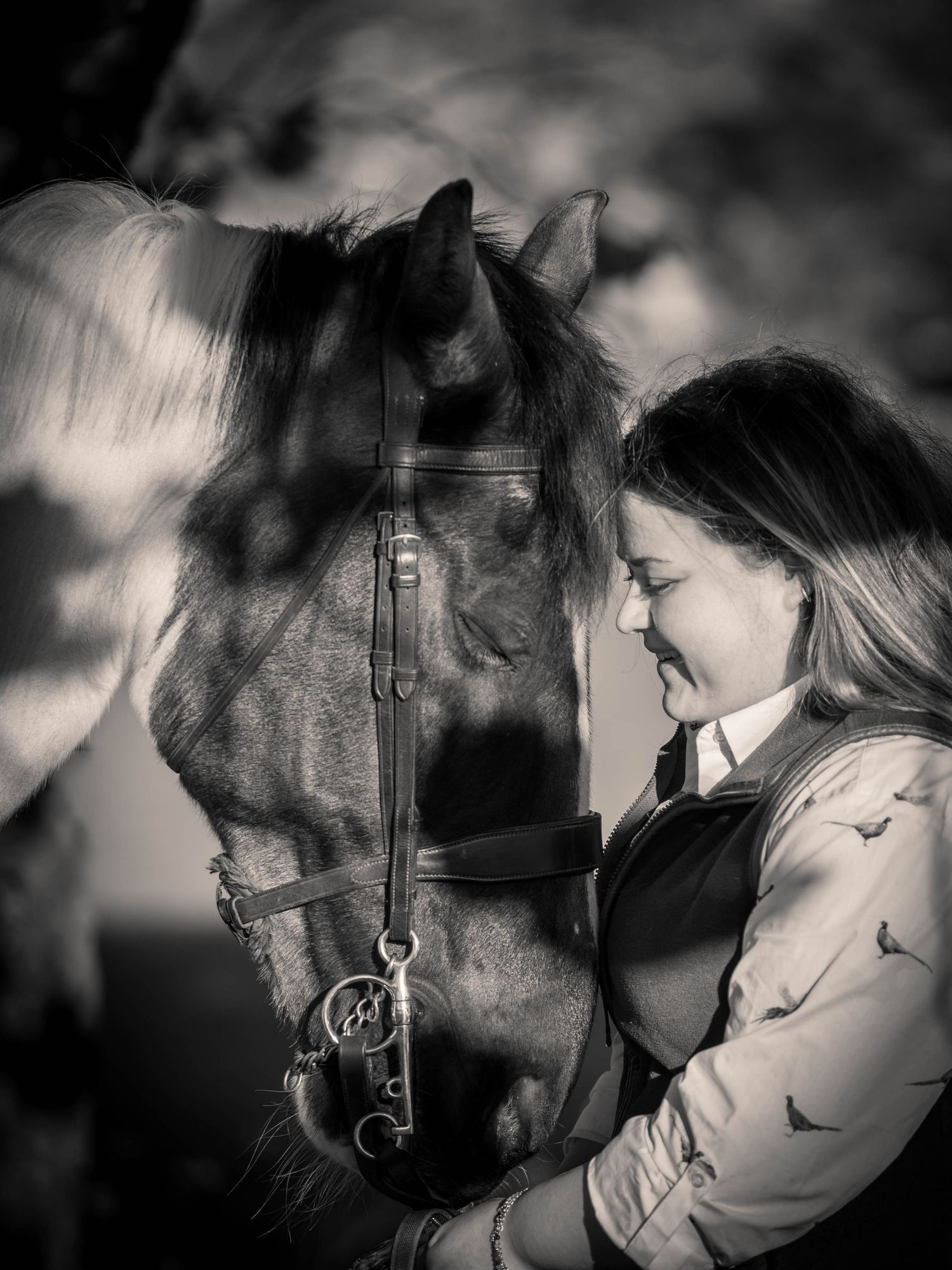 Black and white photo of a smiling young woman in equestrian attire sharing a tender moment with her horse.