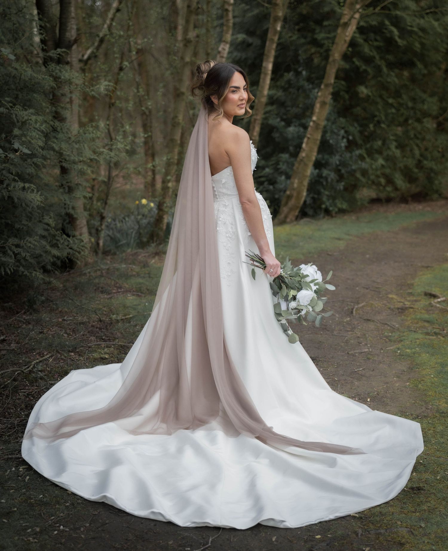 Bride in strapless white gown with long blush veil holding white bouquet, posing outdoors among lush green trees.