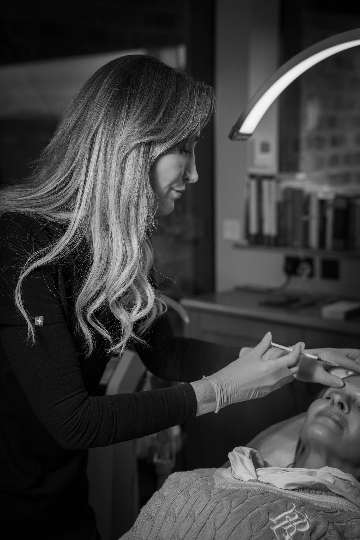 A person in black performs a facial treatment on a client lying on a spa bed in a dimly lit room.