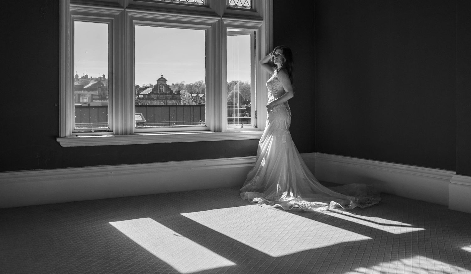Black and white photo of a bride in a wedding gown standing by a sunlit window in an elegant room.