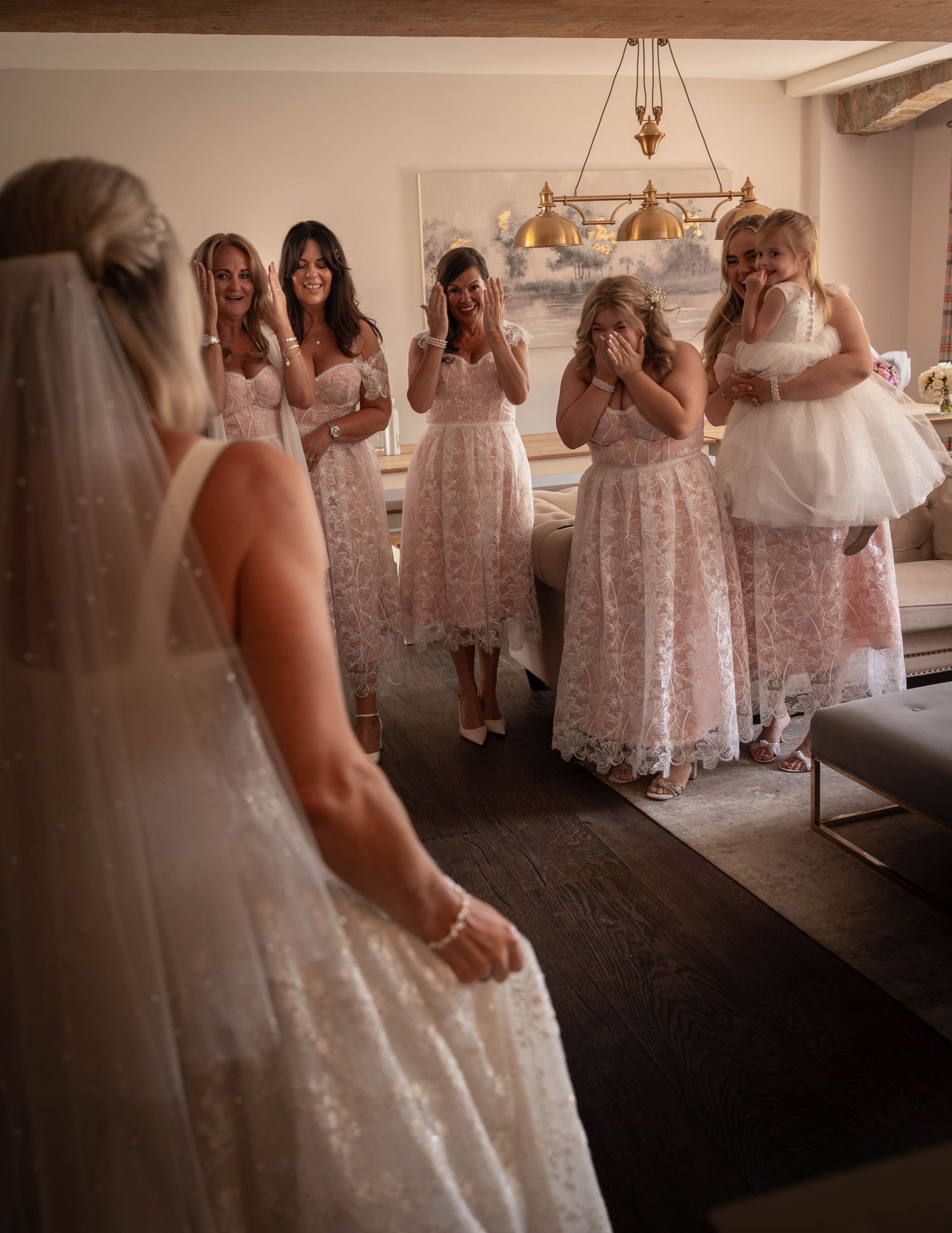 Bride reveals wedding dress to emotional bridesmaids in blush lace gowns, who react with tears and joy in elegant room.