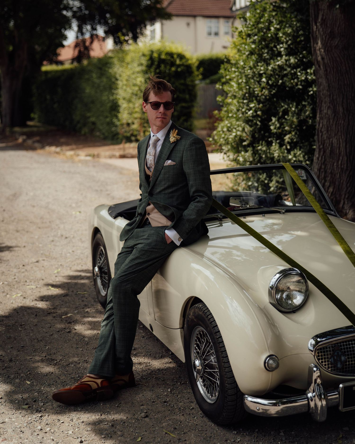 Man in green suit leaning on a cream vintage Austin-Healey sports car on a tree-lined driveway.