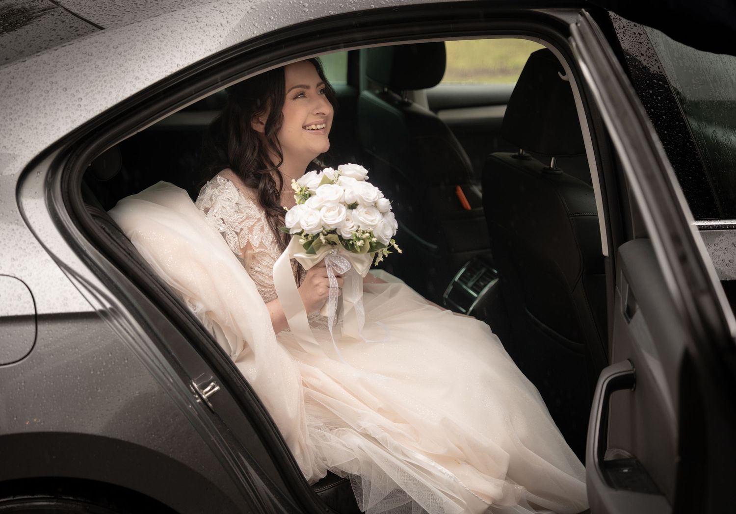Smiling bride in white lace gown holding bouquet seated in car backseat on wedding day.