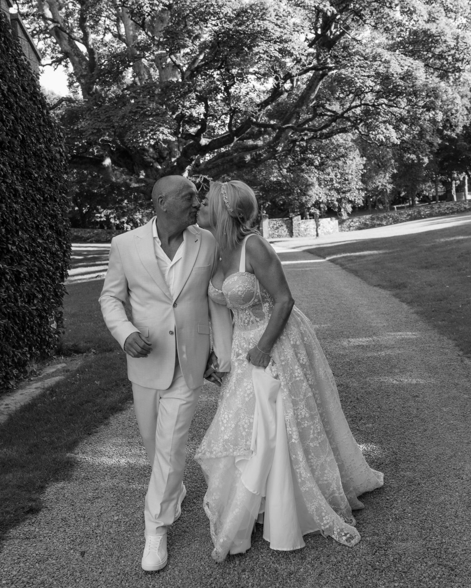 Couple sharing a kiss in wedding attire outdoors, groom in white suit, bride in lace gown, tree-lined path.