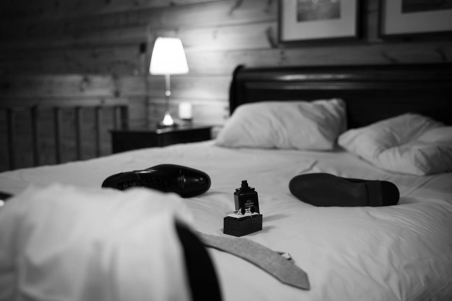Black and white photo of dress shoes, cologne, and accessories on a white hotel bed with lamp in background.