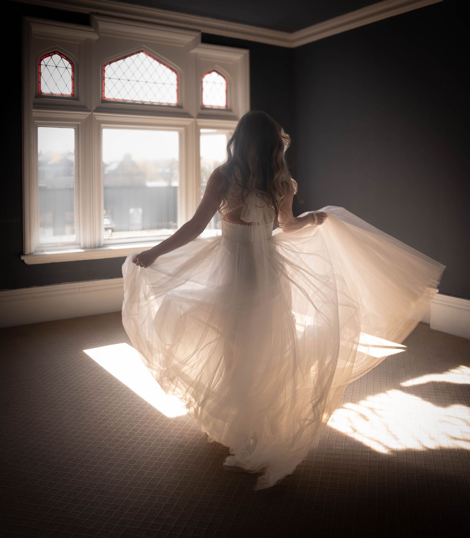 Bride twirling in white wedding gown by sunlit window in elegant room.
