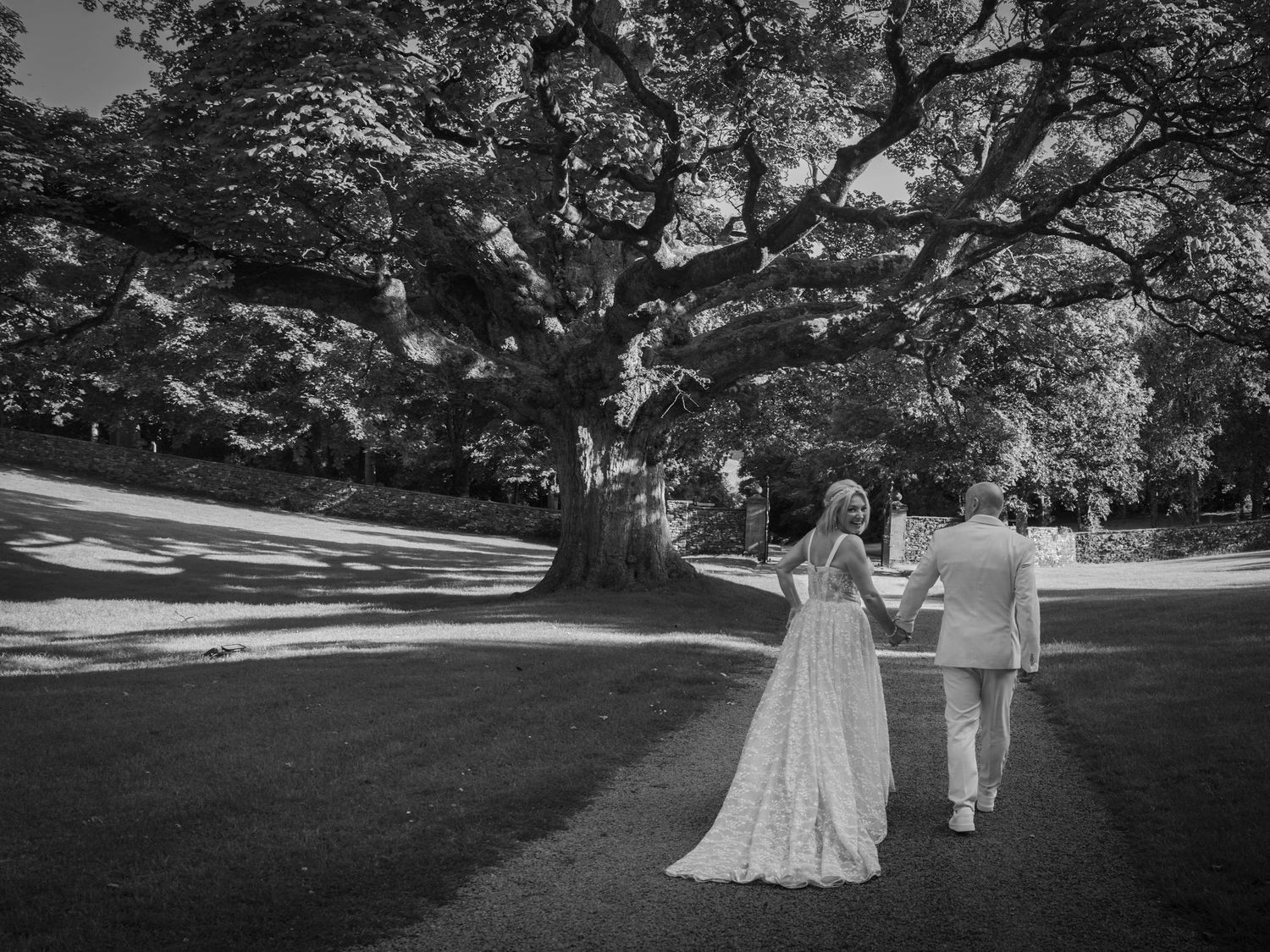 Black and white photo of a bride and groom walking hand in hand beneath a large oak tree on a sunny day.