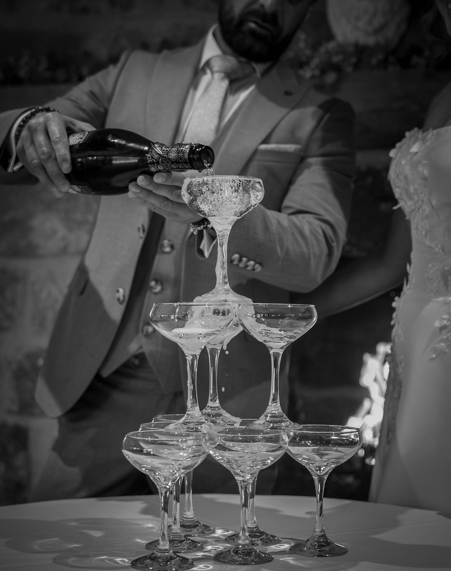 Black and white photo of a man in a suit pouring champagne into a pyramid of coupe glasses at a wedding reception.