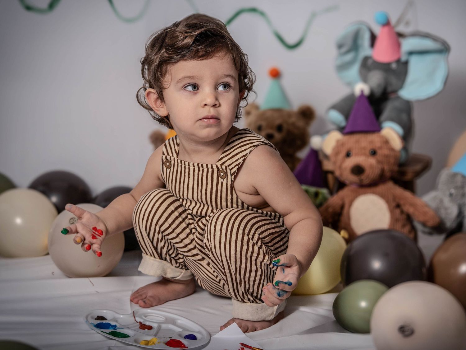 Child in brown striped dress lies on bed surrounded by colorful stuffed animals and toys in playful setting.