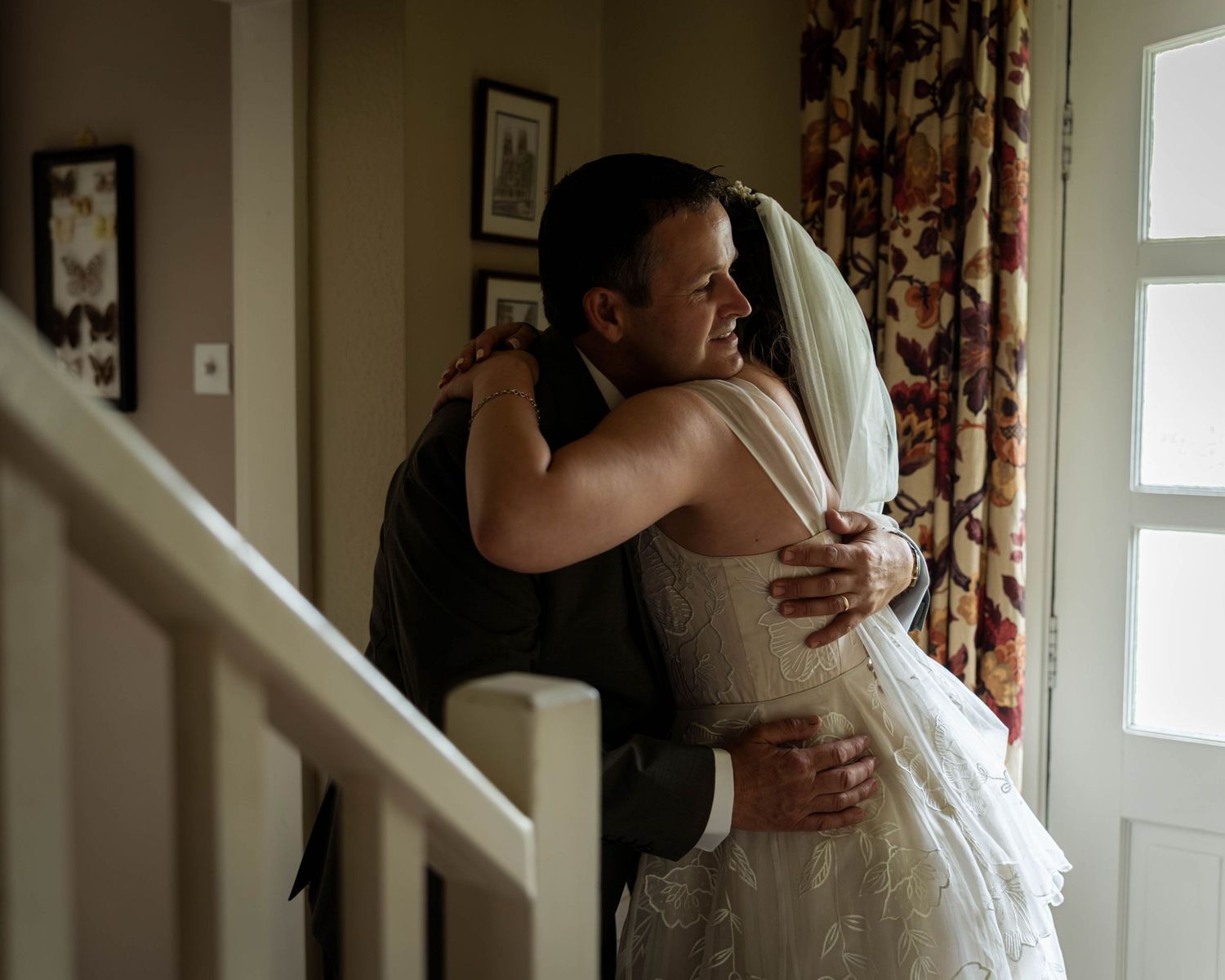 Bride in white lace gown and veil shares an emotional hug with a guest in a warmly lit home interior.