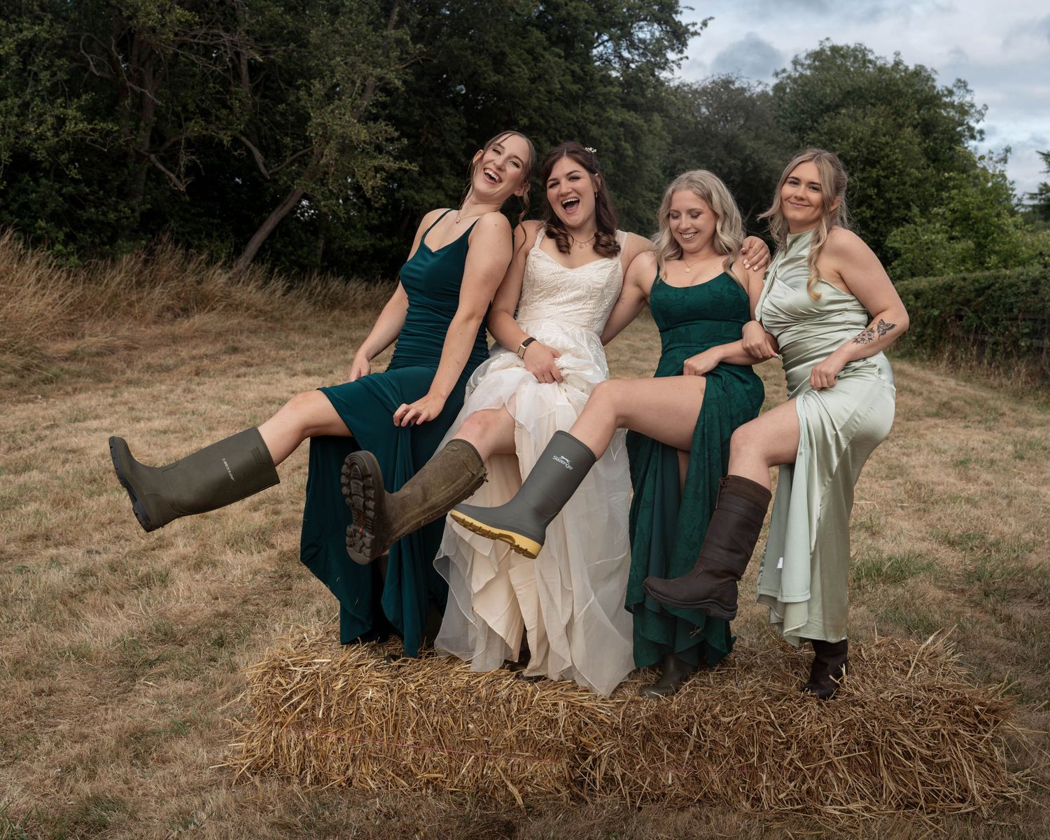 Bride and bridesmaids in green dresses and rain boots sitting on hay bales outdoors at a rustic countryside wedding.