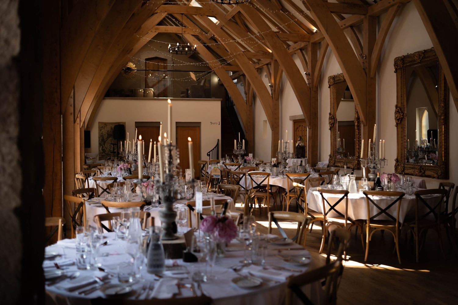 Elegant barn wedding reception with wooden beams, candlelit tables, and rustic cross-back chairs set for a formal dinner.