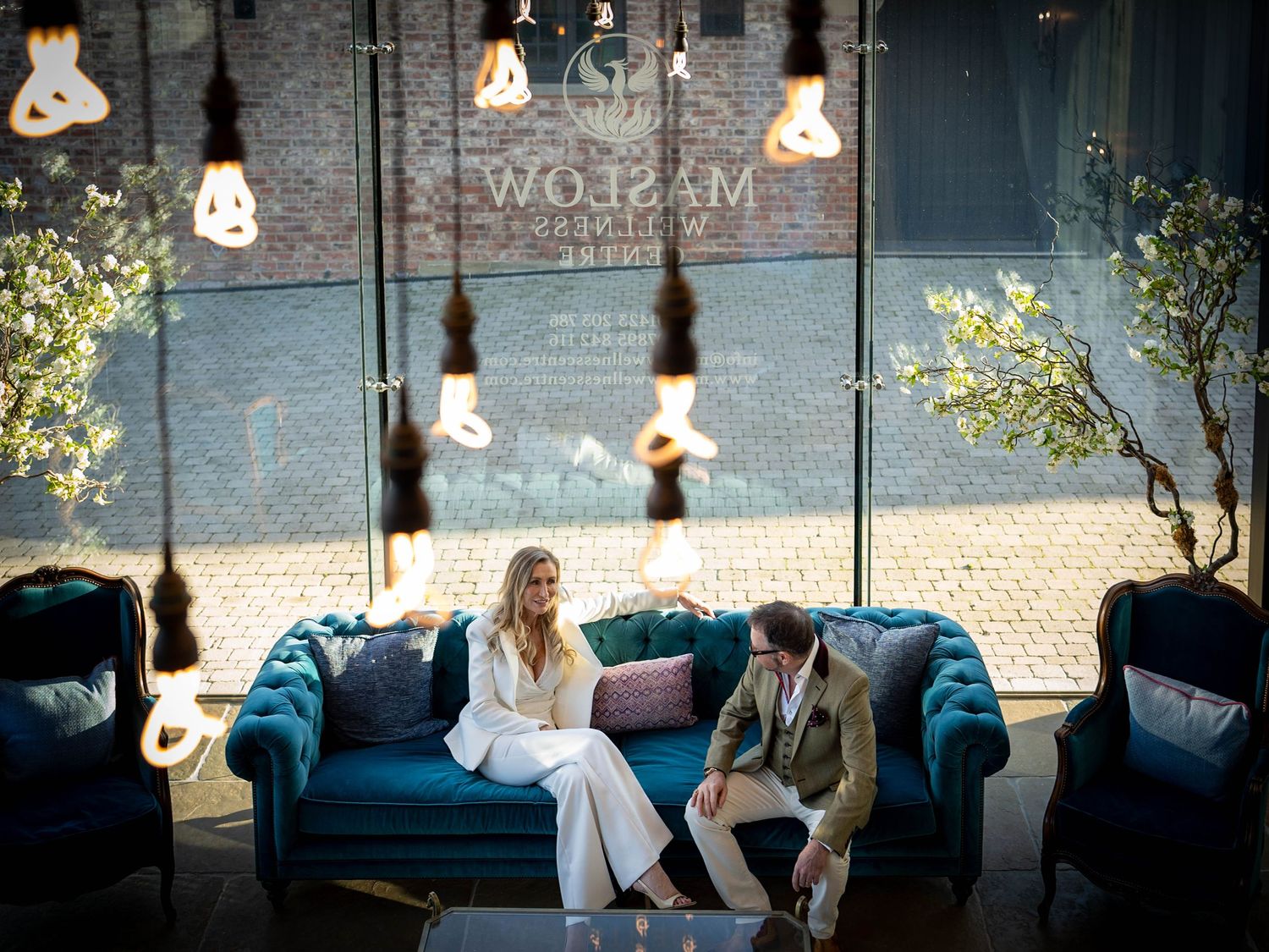 Couple sitting on teal velvet sofa under Edison bulbs in stylish hotel lobby with brick backdrop.
