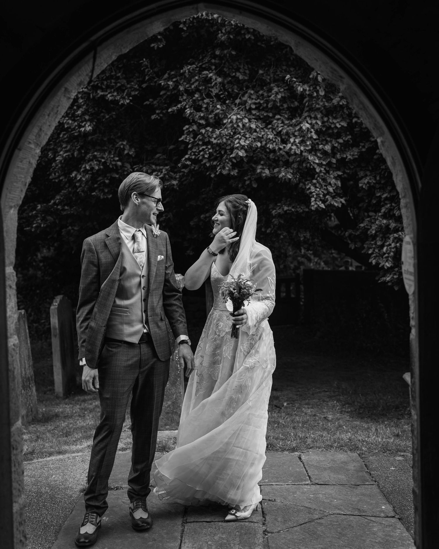 Black and white wedding photo of bride and groom sharing a tender moment under a stone arch doorway.