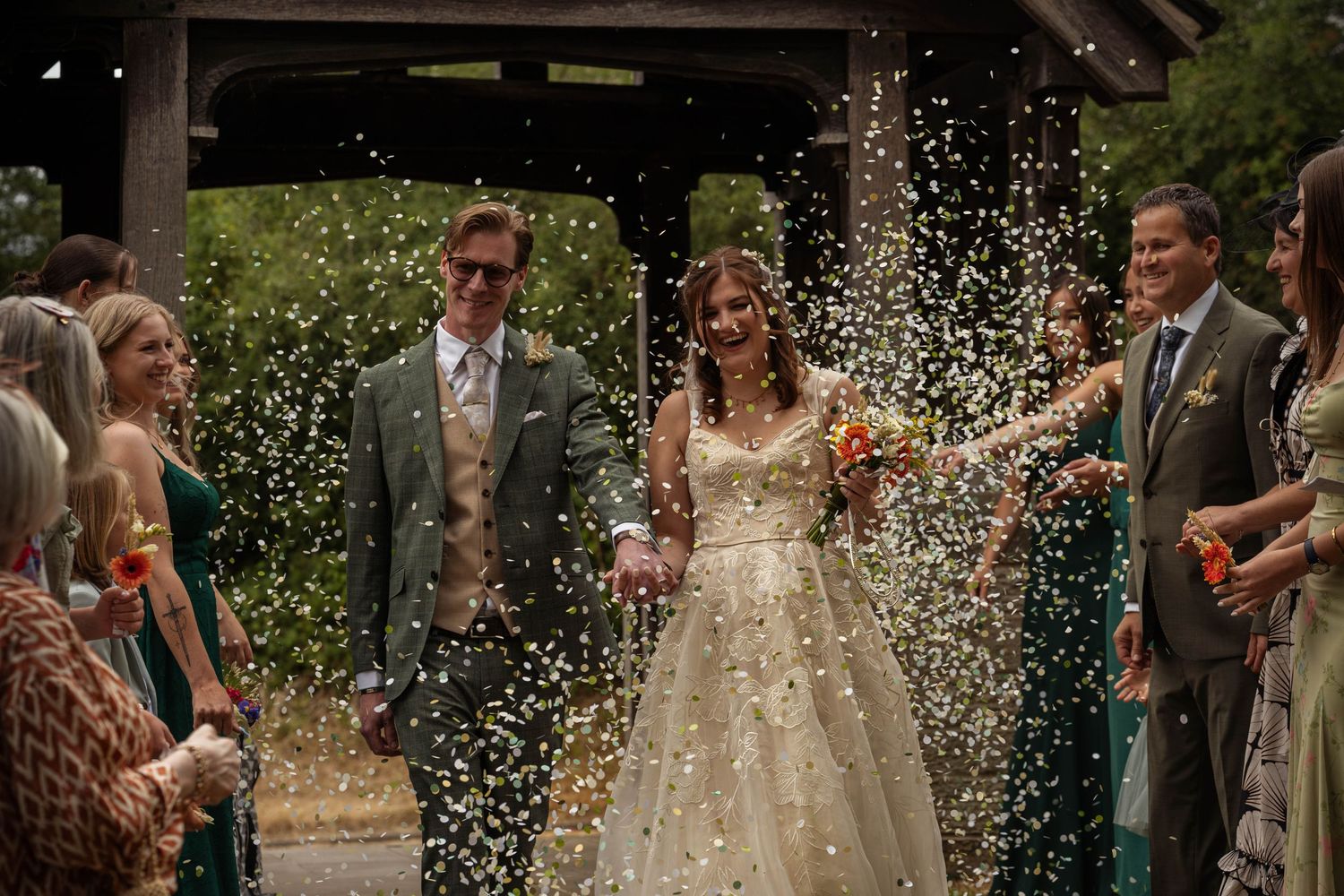 Bride and groom walk through confetti shower at outdoor wedding ceremony, surrounded by smiling guests.