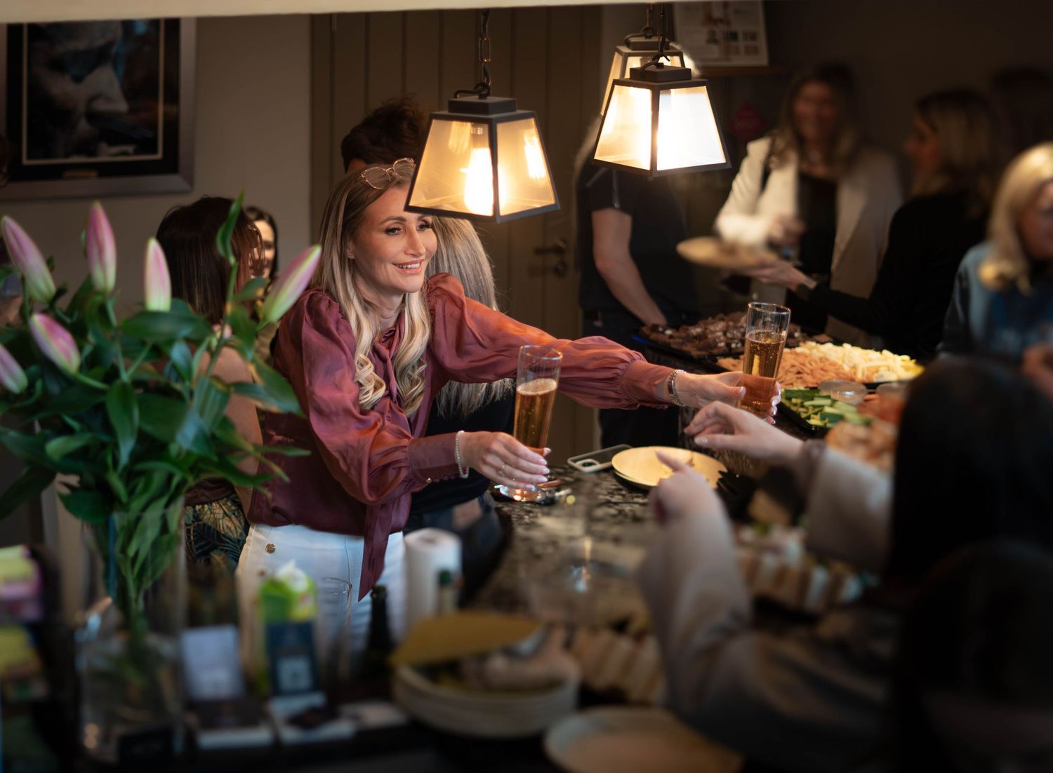 Women socializing at a warm dinner party, clinking glasses over a table with food and flowers under soft lamp light.