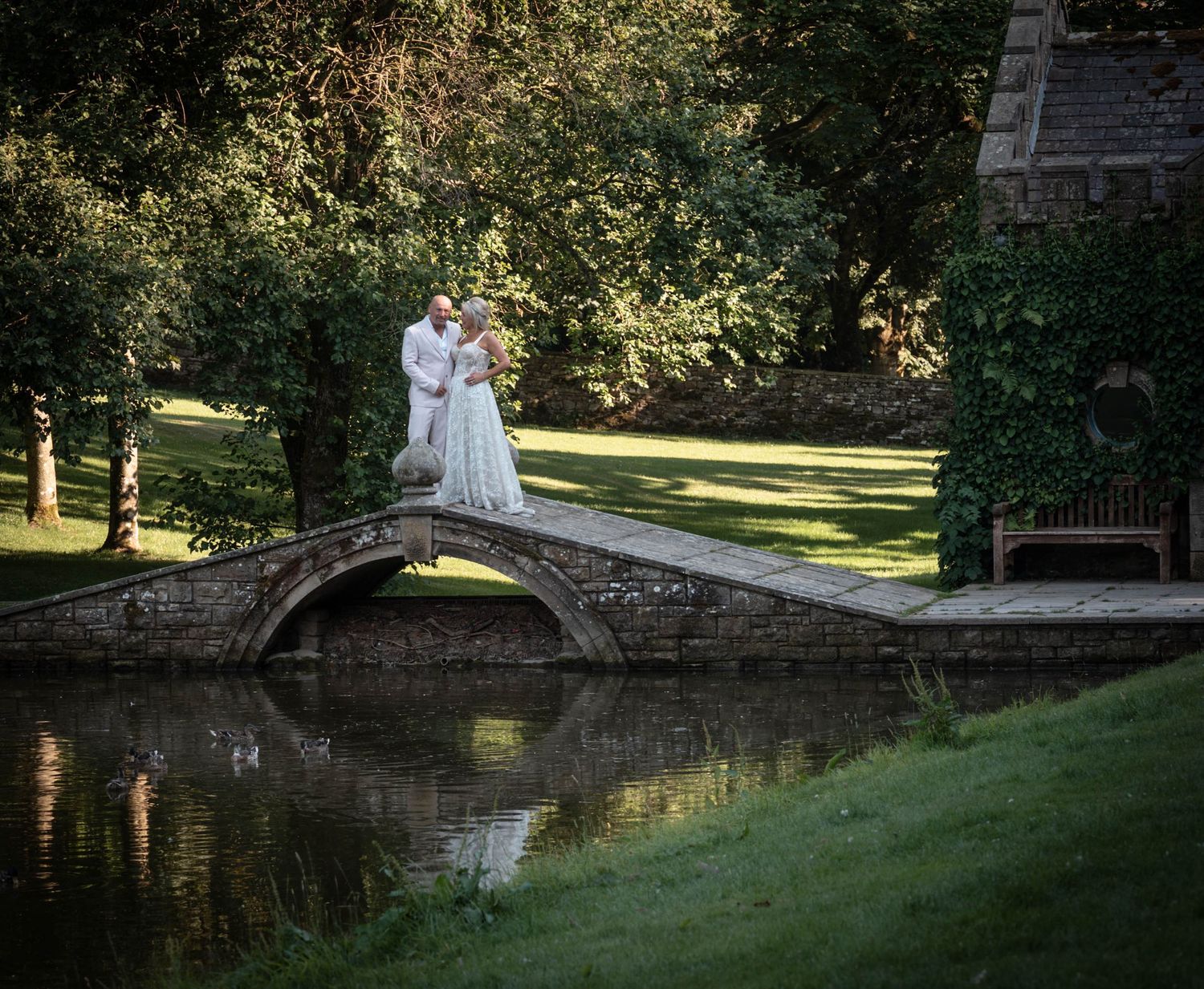 Bride and groom share a romantic kiss on a stone arch bridge over a calm pond in a lush green garden.