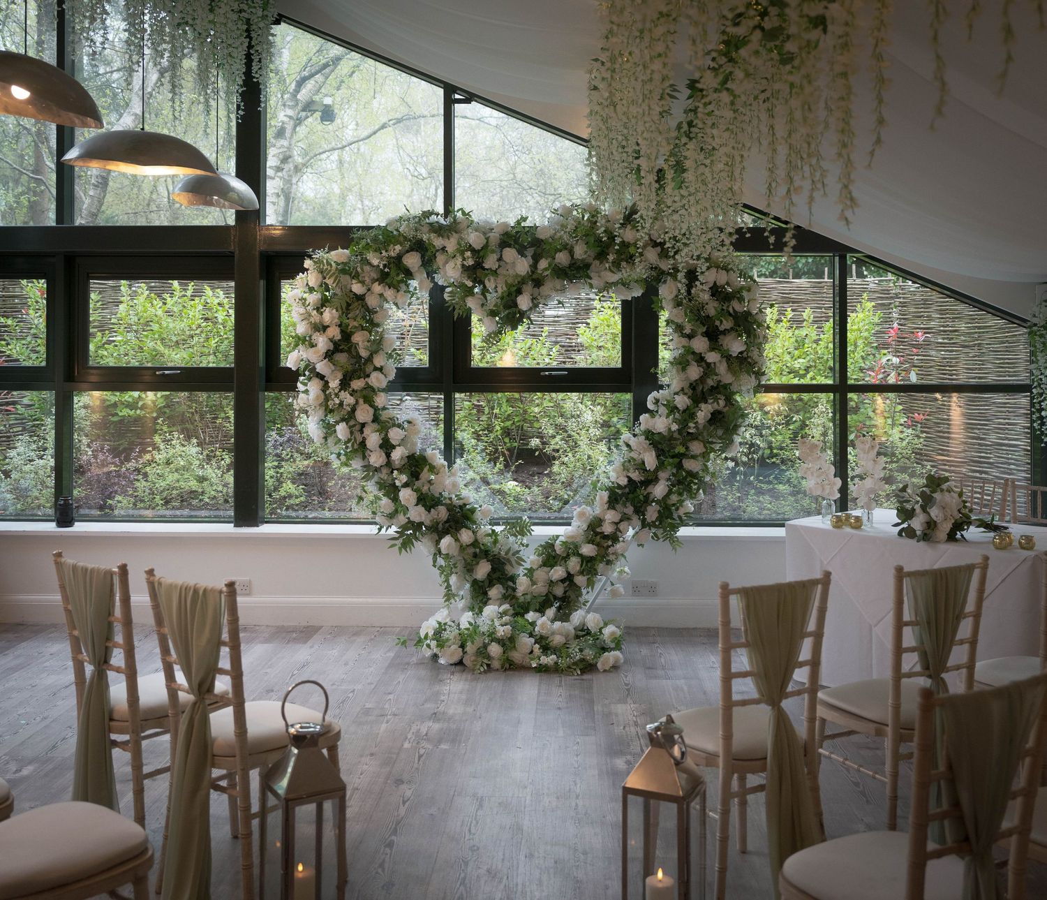 Elegant wedding ceremony setup with heart-shaped floral arch, chiavari chairs, and hanging greenery in a bright venue.