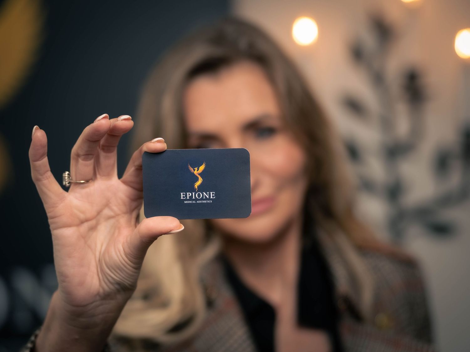 Someone holds up a dark colored business card with a logo against a blurred background with bokeh lights.
