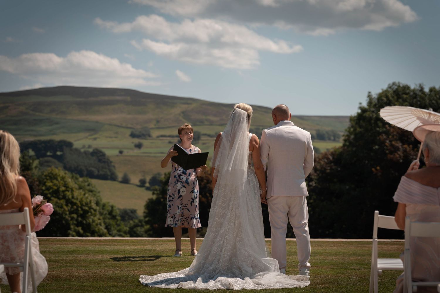 Outdoor wedding ceremony with bride and groom before officiant, rolling green hills and blue sky backdrop.