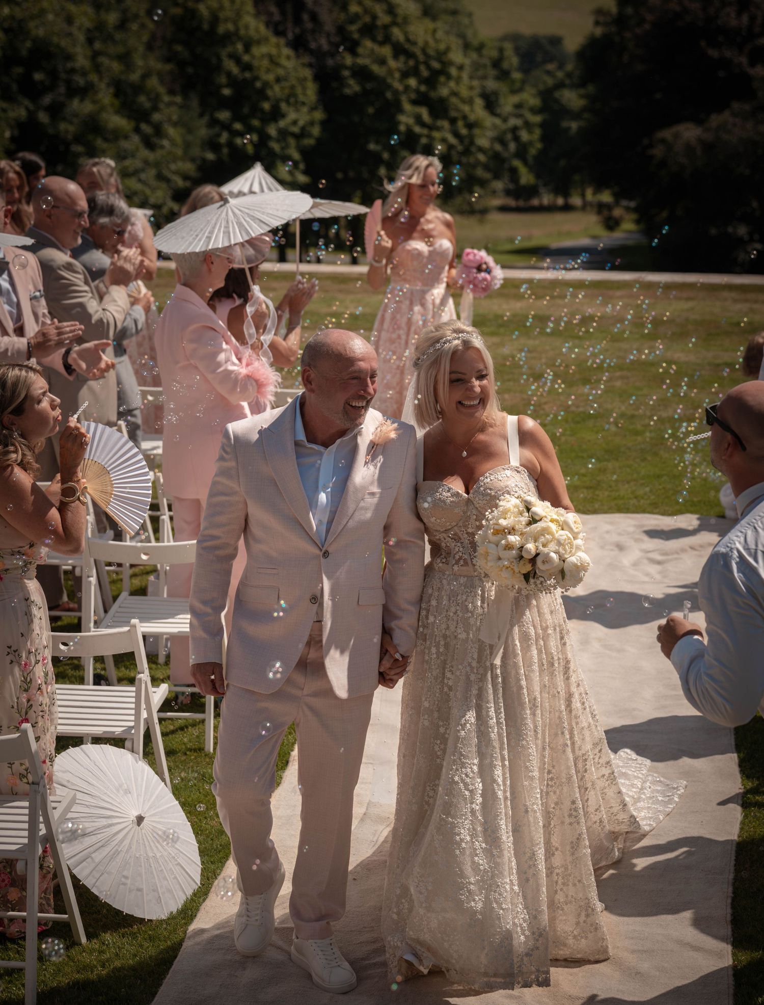 Bride and groom walk down outdoor aisle holding hands, surrounded by bubbles and guests on a sunny wedding day.