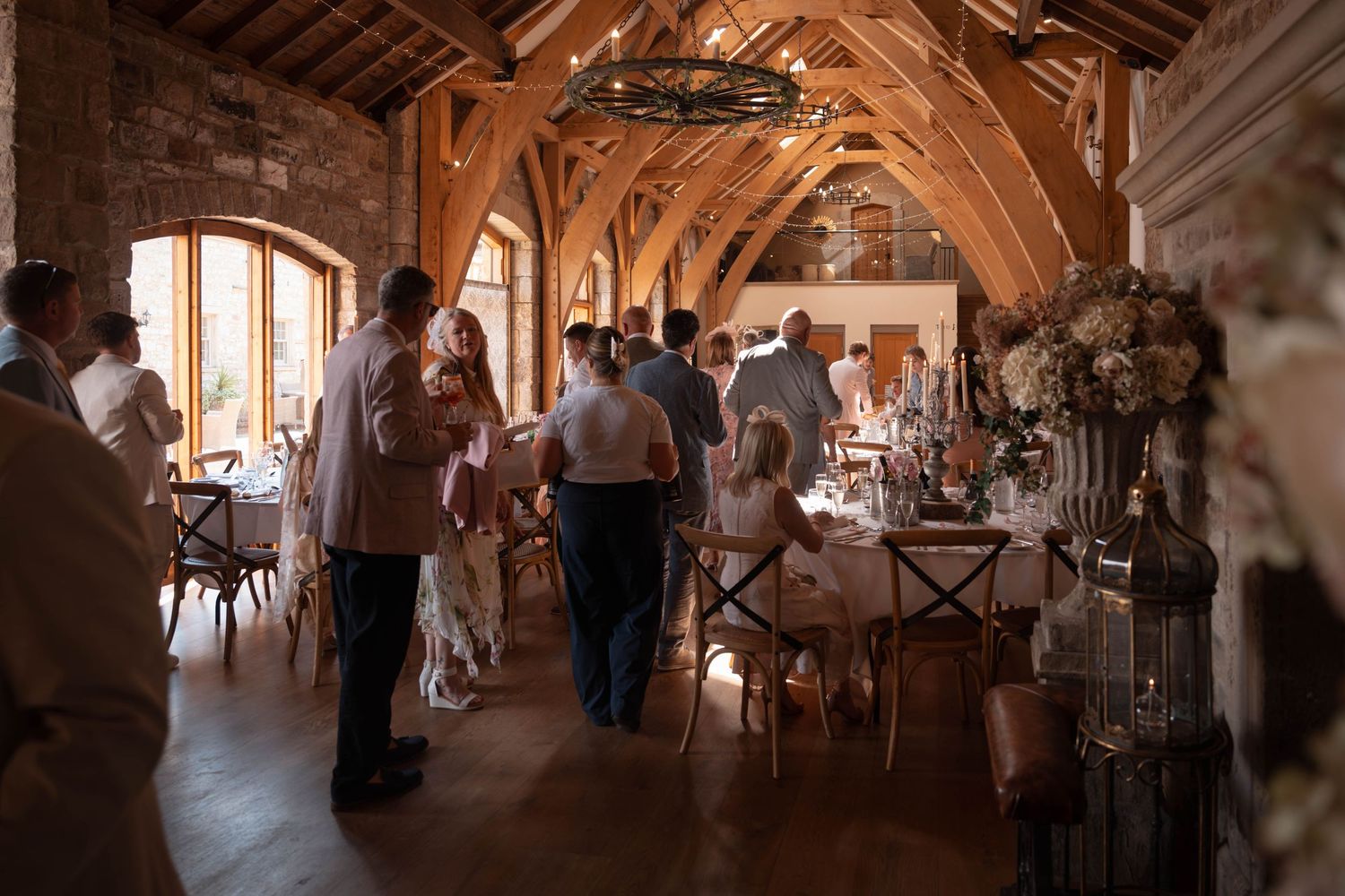 Guests mingle in a rustic barn wedding venue with wooden beams, fairy lights, and elegant floral decorations.