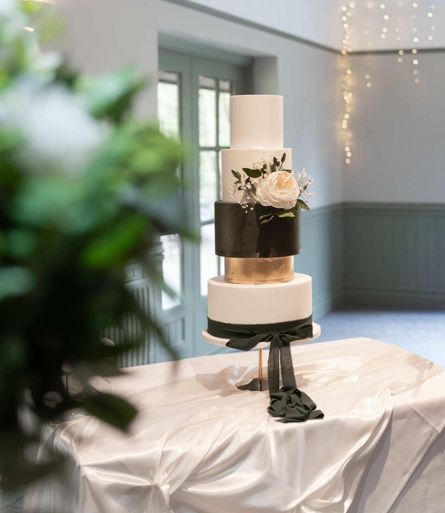 Elegant four-tier wedding cake with black, gold, and white layers adorned with white roses on a draped table.
