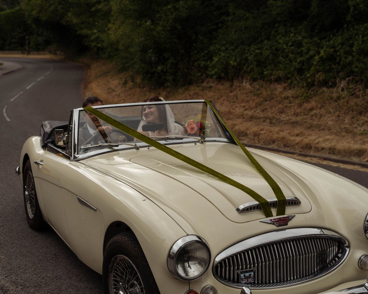 Vintage cream Austin-Healey convertible decorated with green ribbon, carrying a couple on a scenic country road.