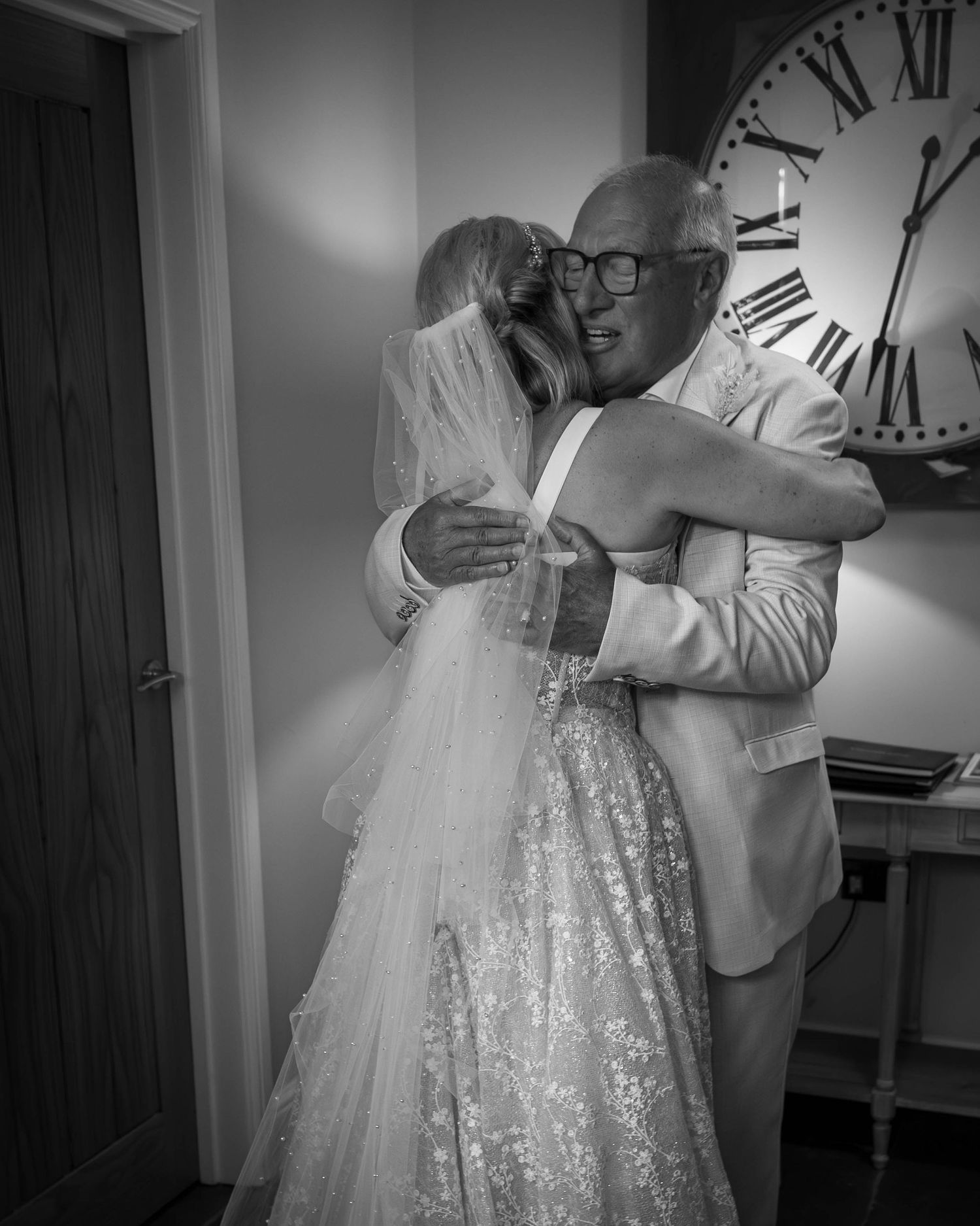 Black and white photo of a bride in lace gown embracing a man in suit, large clock on wall behind them.