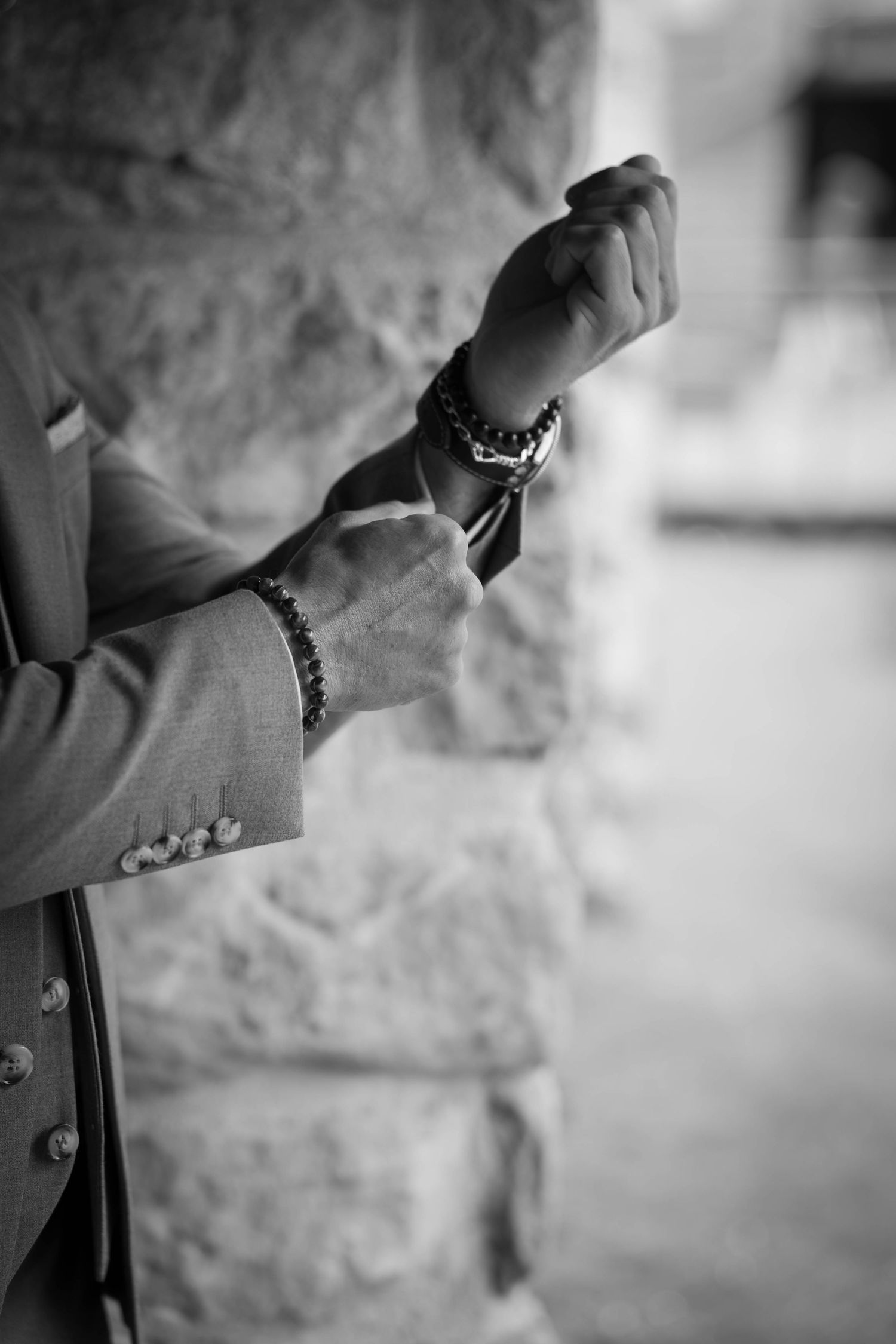Black and white photo of a man in a suit adjusting his cufflinks, wearing multiple beaded bracelets.
