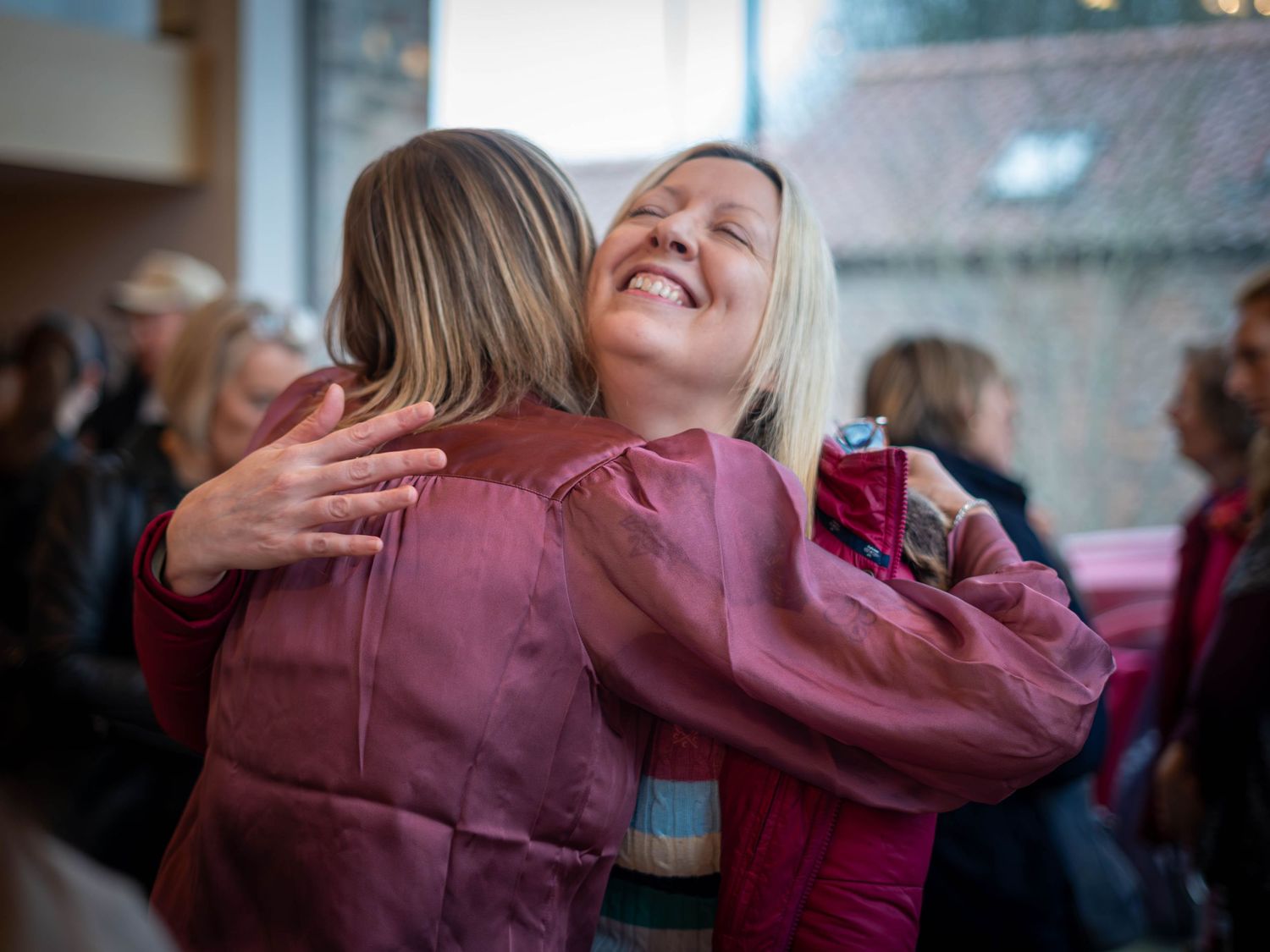 Two women sharing a warm hug and smiling at an indoor social gathering, both wearing pink and mauve outfits.