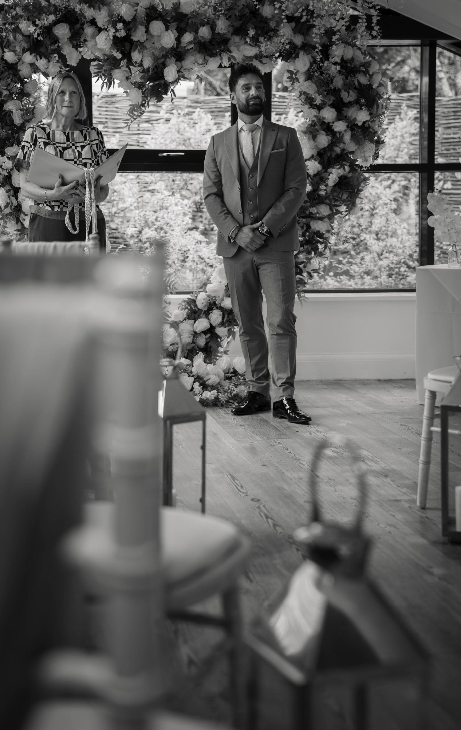 Groom in grey suit stands at wedding altar adorned with floral arch and wreath in elegant venue.