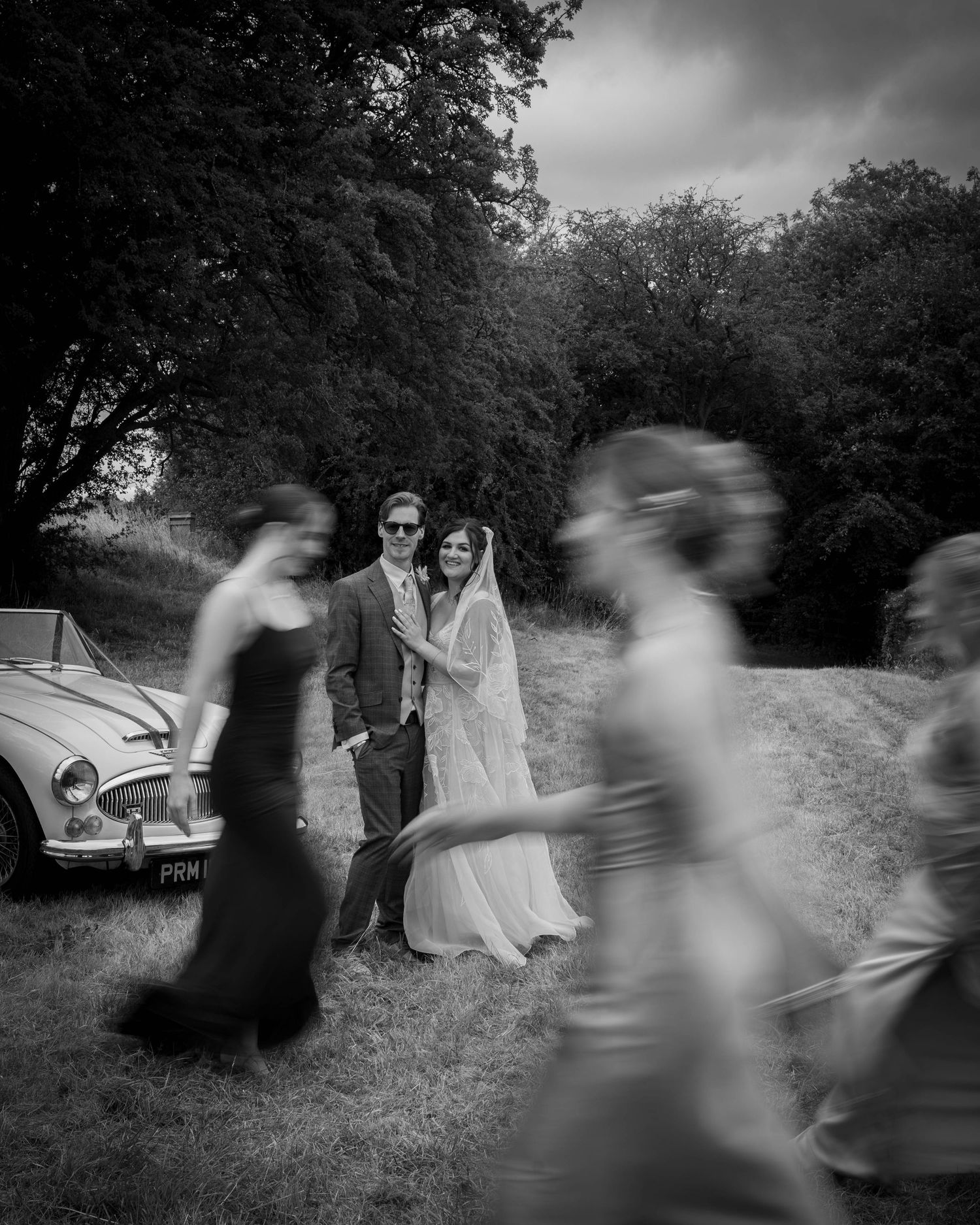 Black and white wedding photo of bride and groom posing by vintage car while bridesmaids walk past in motion blur.
