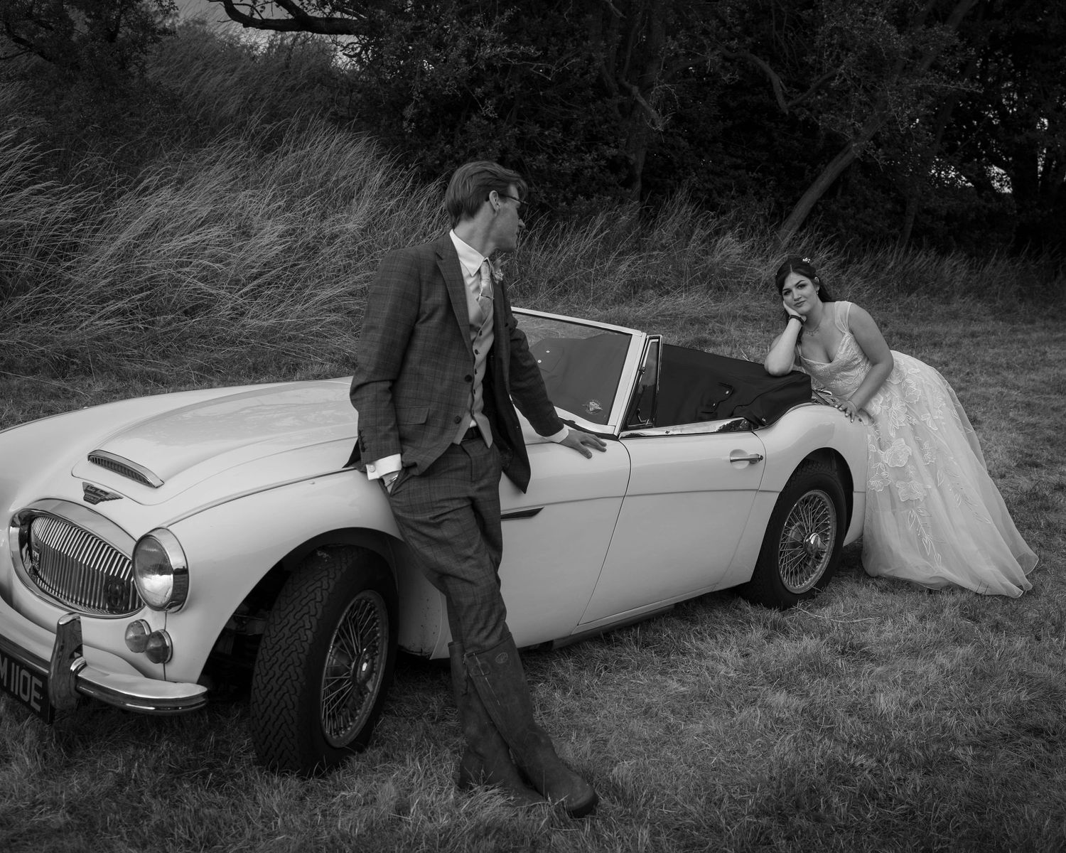 Black and white photo of a bride and groom posing elegantly beside a classic vintage convertible car outdoors.