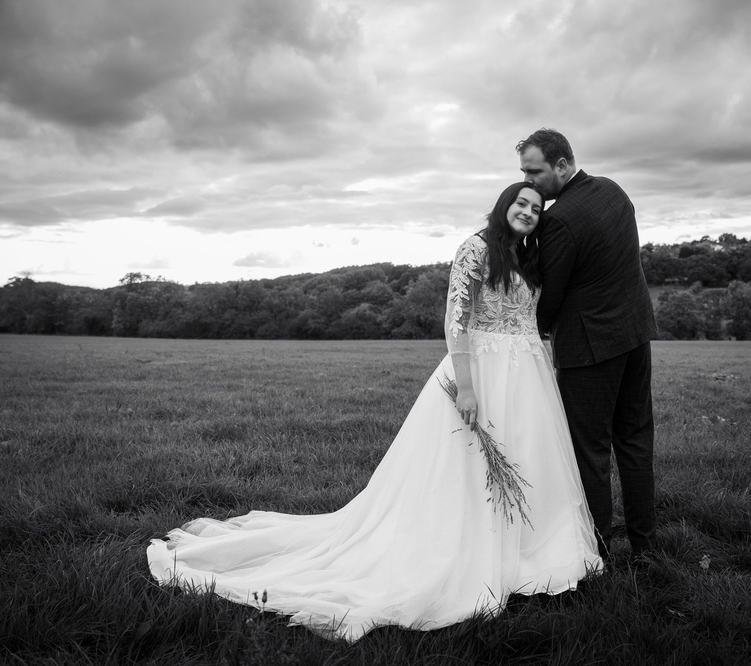 A romantic black and white wedding photo series showing a couple embracing in a windswept meadow with dramatic clouds.