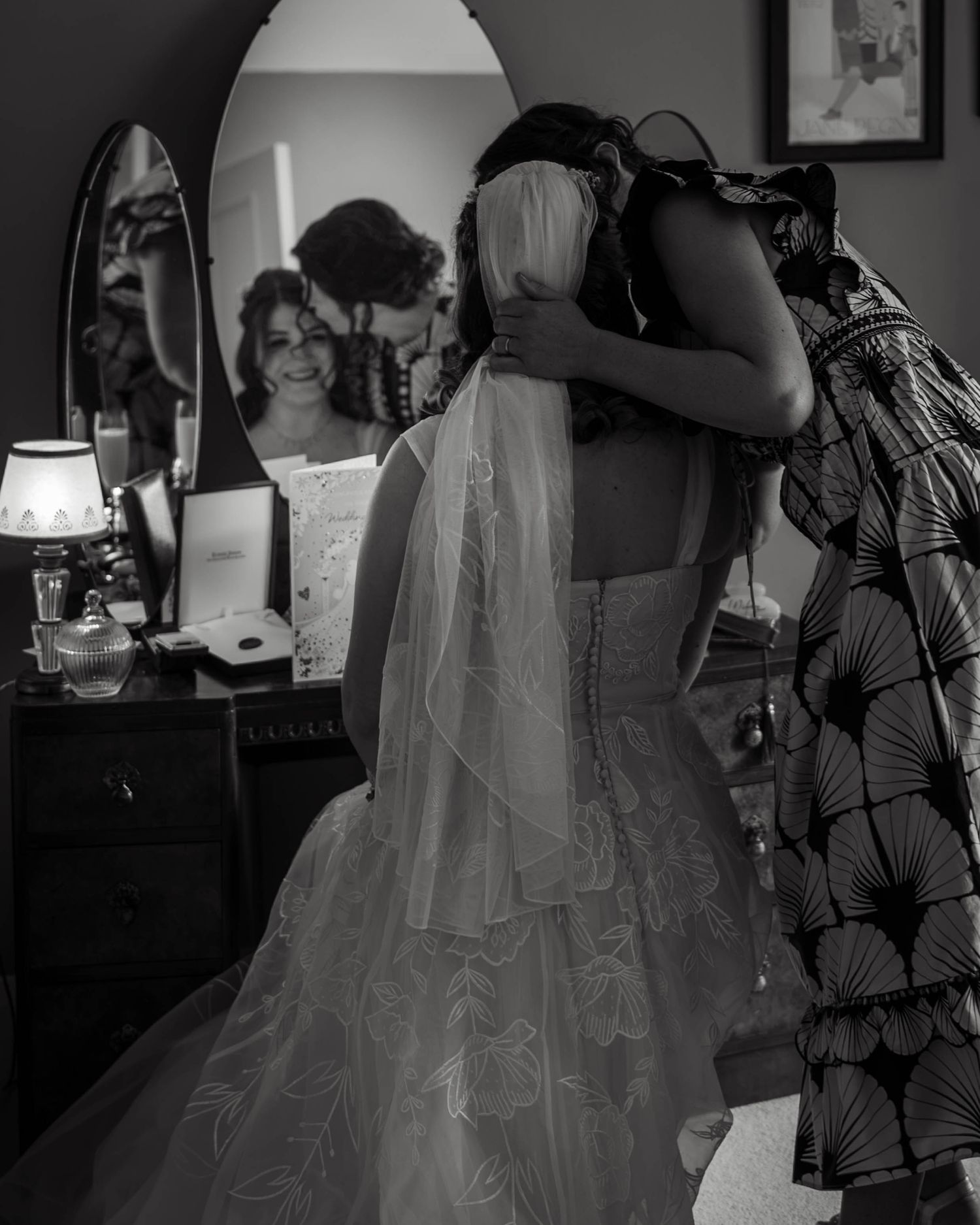 Black and white photo of a bride in lace gown embracing bridesmaid, mirror reflection showing her smile on wedding day.