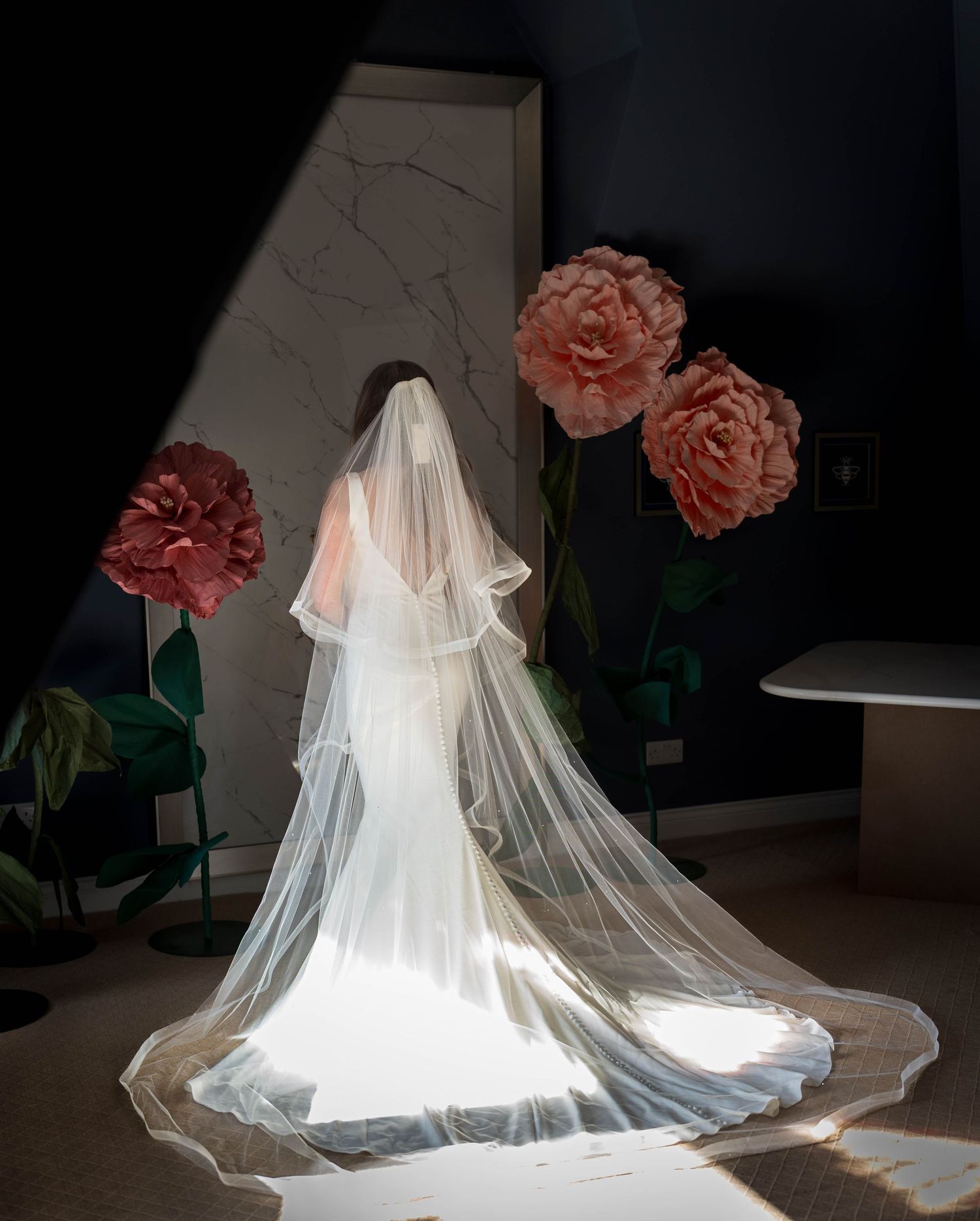 Bride in white gown and long veil stands in dramatic light surrounded by large pink peony decorations.