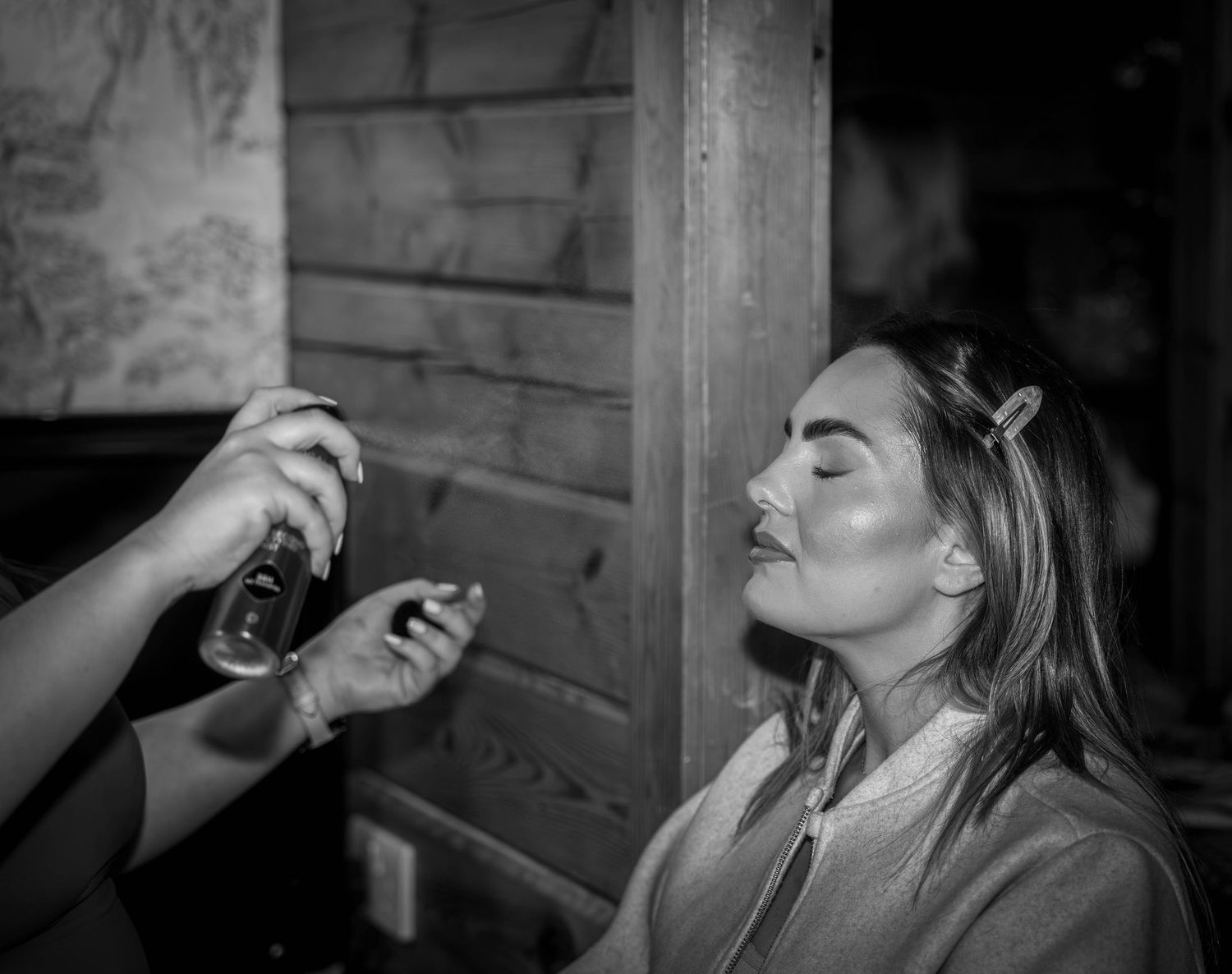 Black and white photo of a woman getting makeup applied with a spray bottle in a rustic wooden setting.