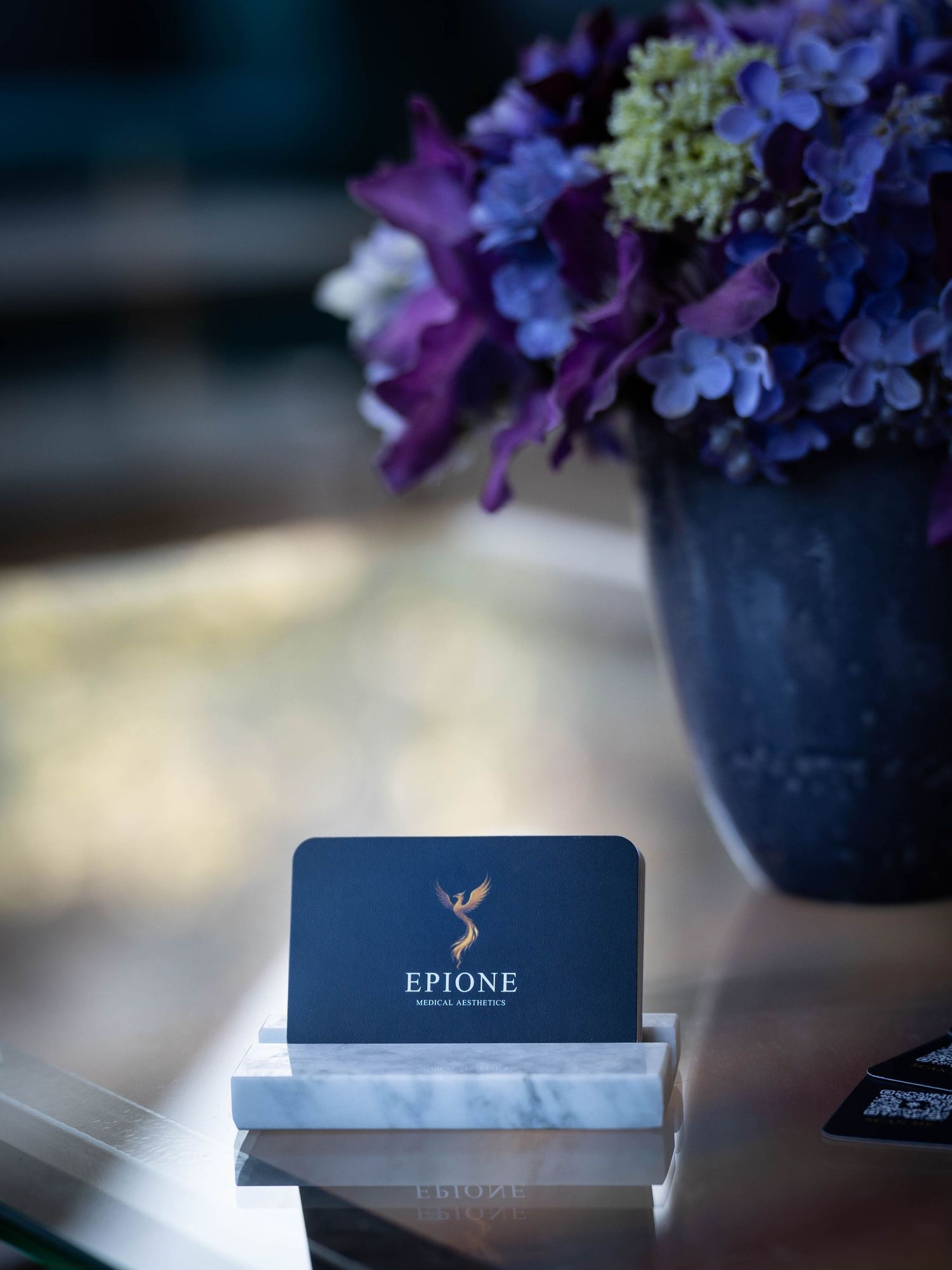 Purple hydrangea flowers arranged in a navy blue vase next to an Epione business card holder on a light surface.