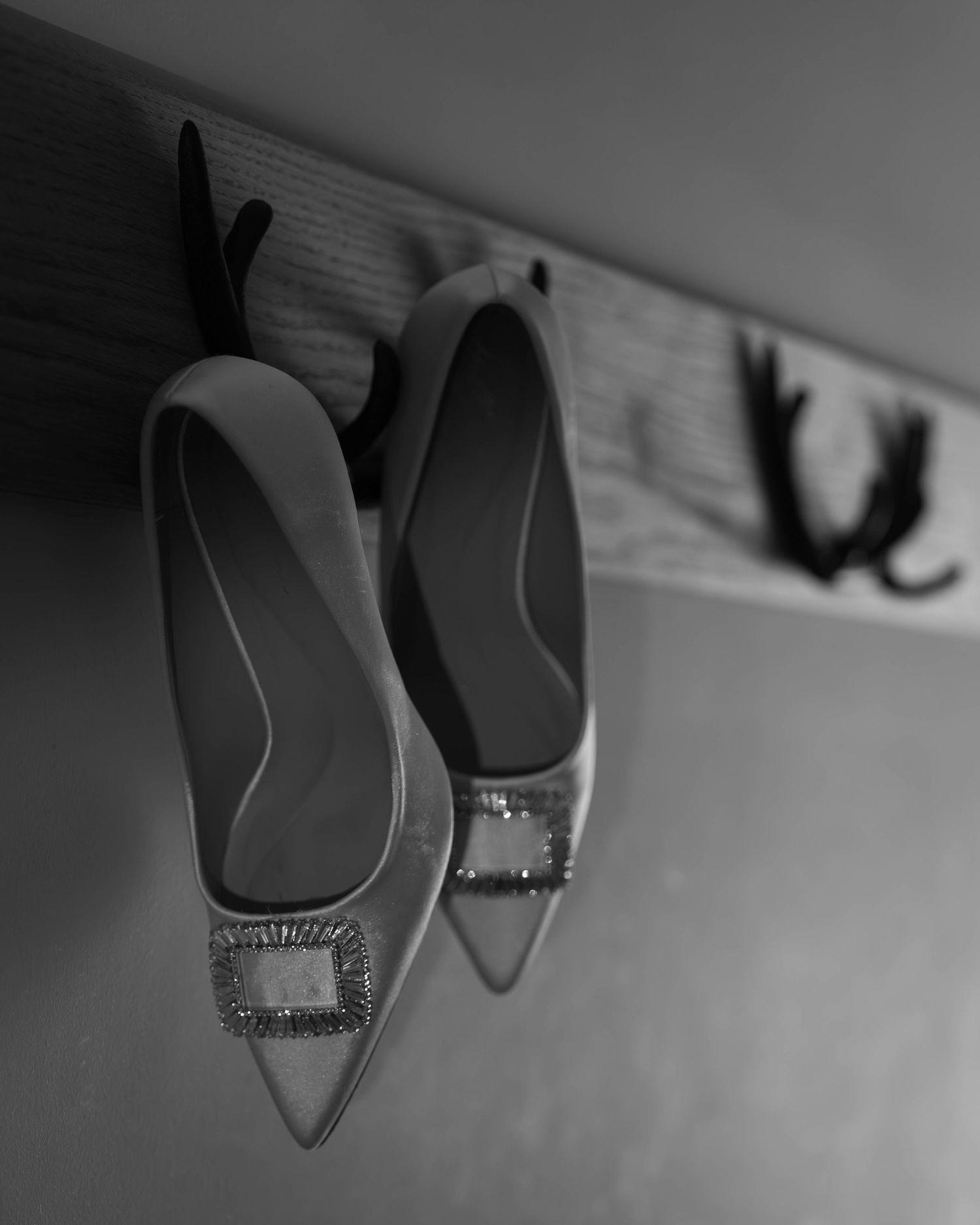 Black and white photo of elegant pointed-toe heels with jeweled buckles hanging on a wooden wall hook.