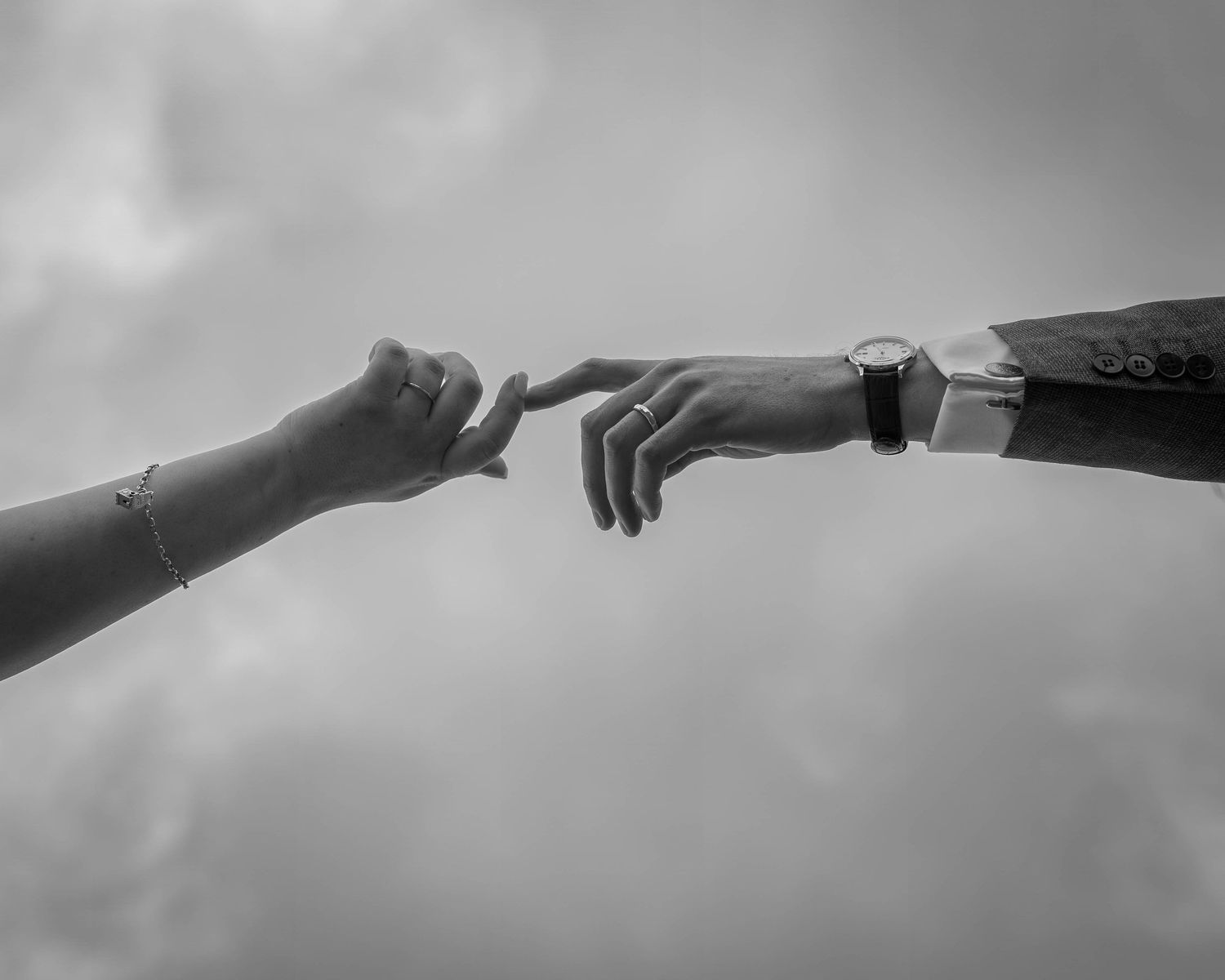 Black and white photo of two hands almost touching, one wearing a bracelet, the other a watch and suit jacket.
