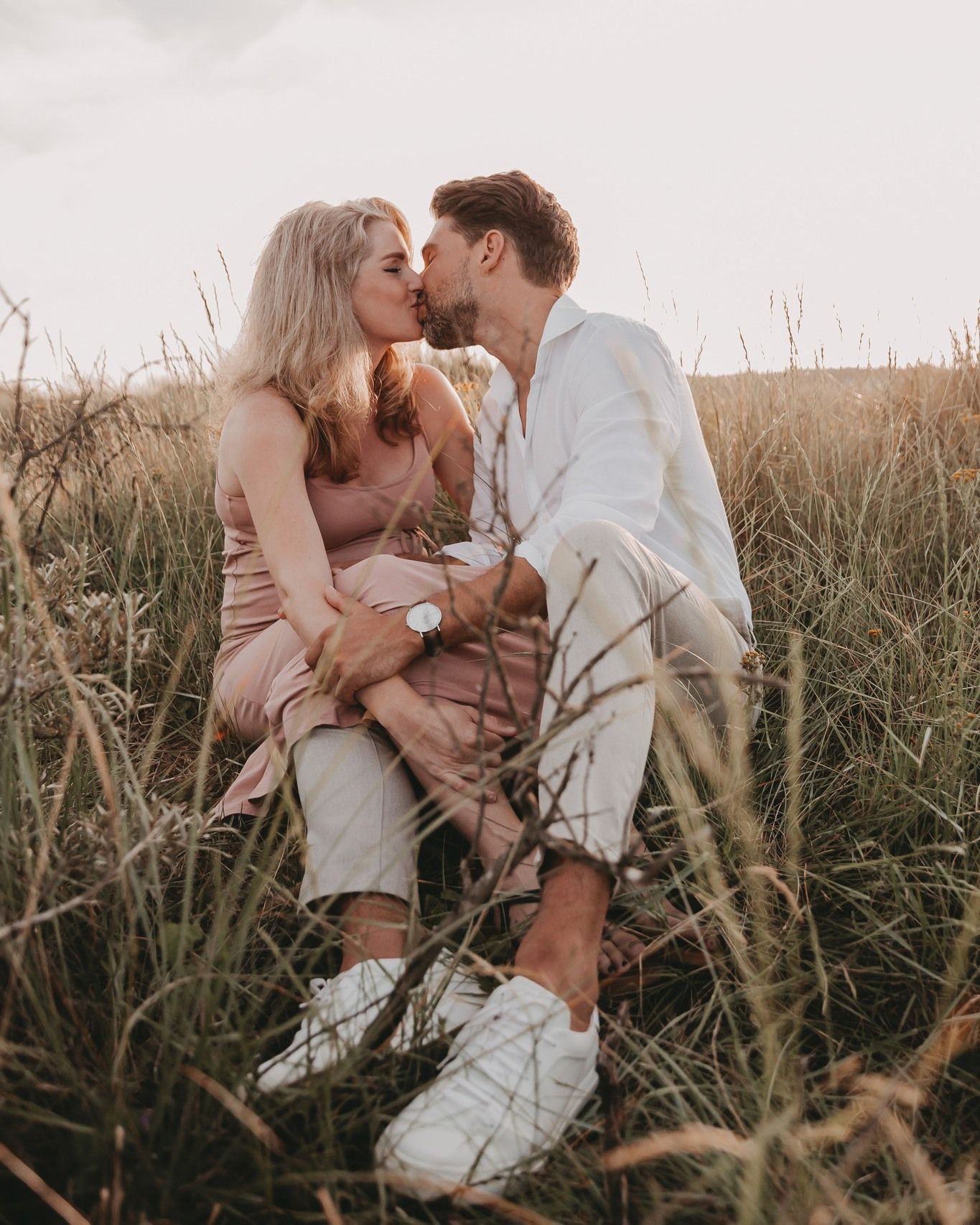 Koppel geeft elkaar een kus tijdens een fotoshoot in duinen tijdens zonsondergang.