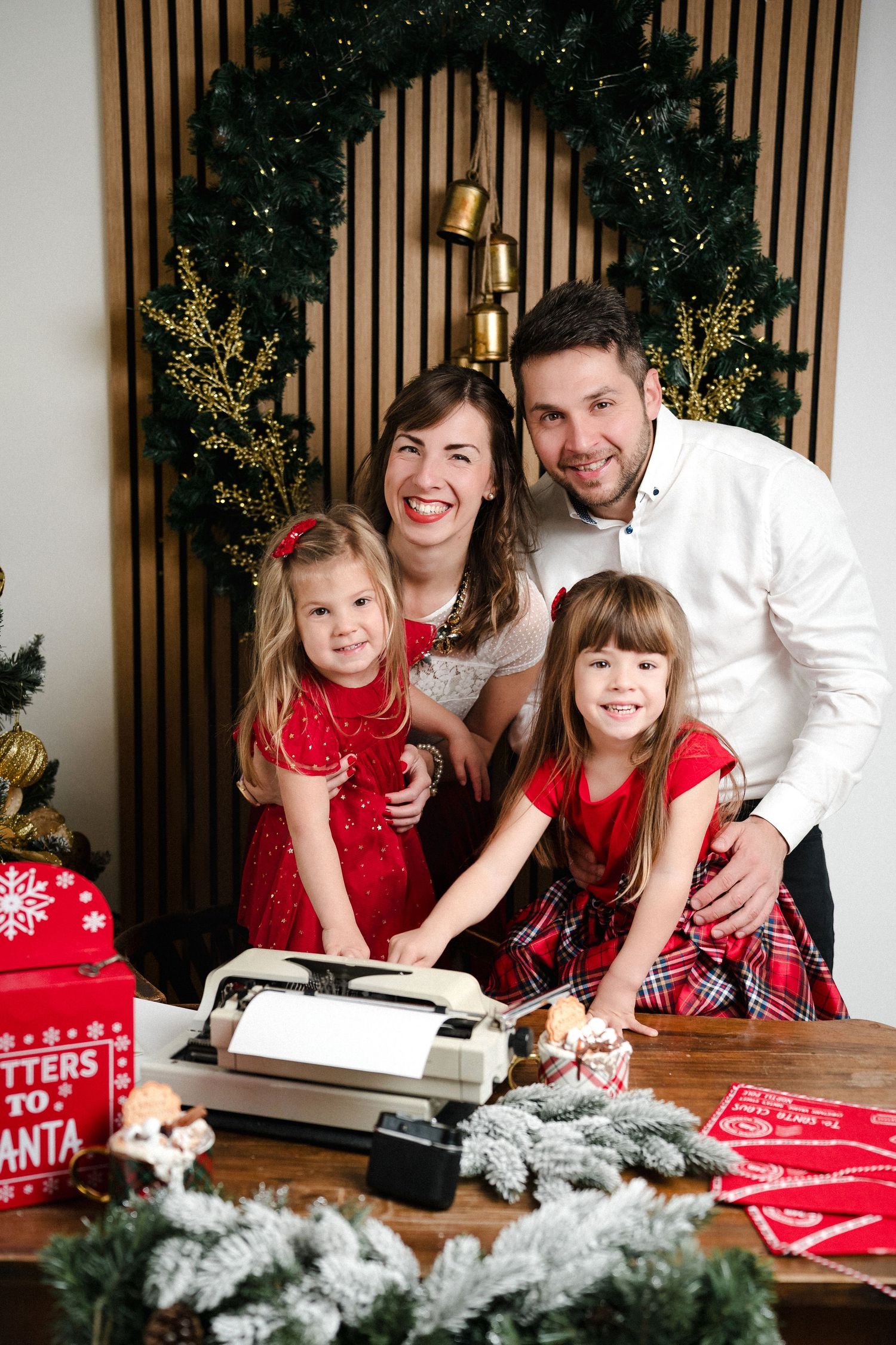 Family posing with typewriter and Letters to Santa box in festive Christmas decorated room with wreath and holiday gifts.