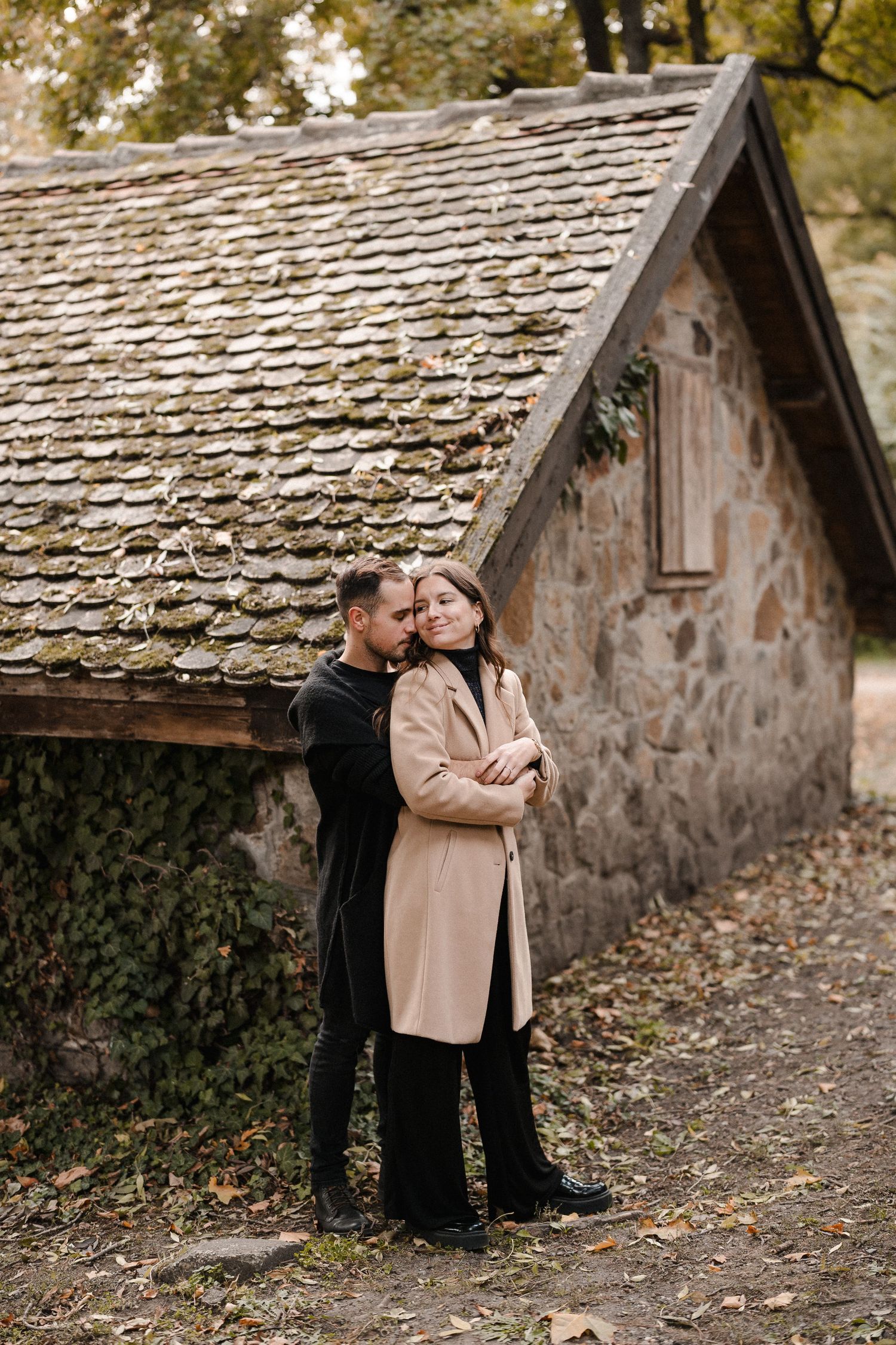Romantic couple sitting against rustic stone building with wooden shingle roof in autumn setting.