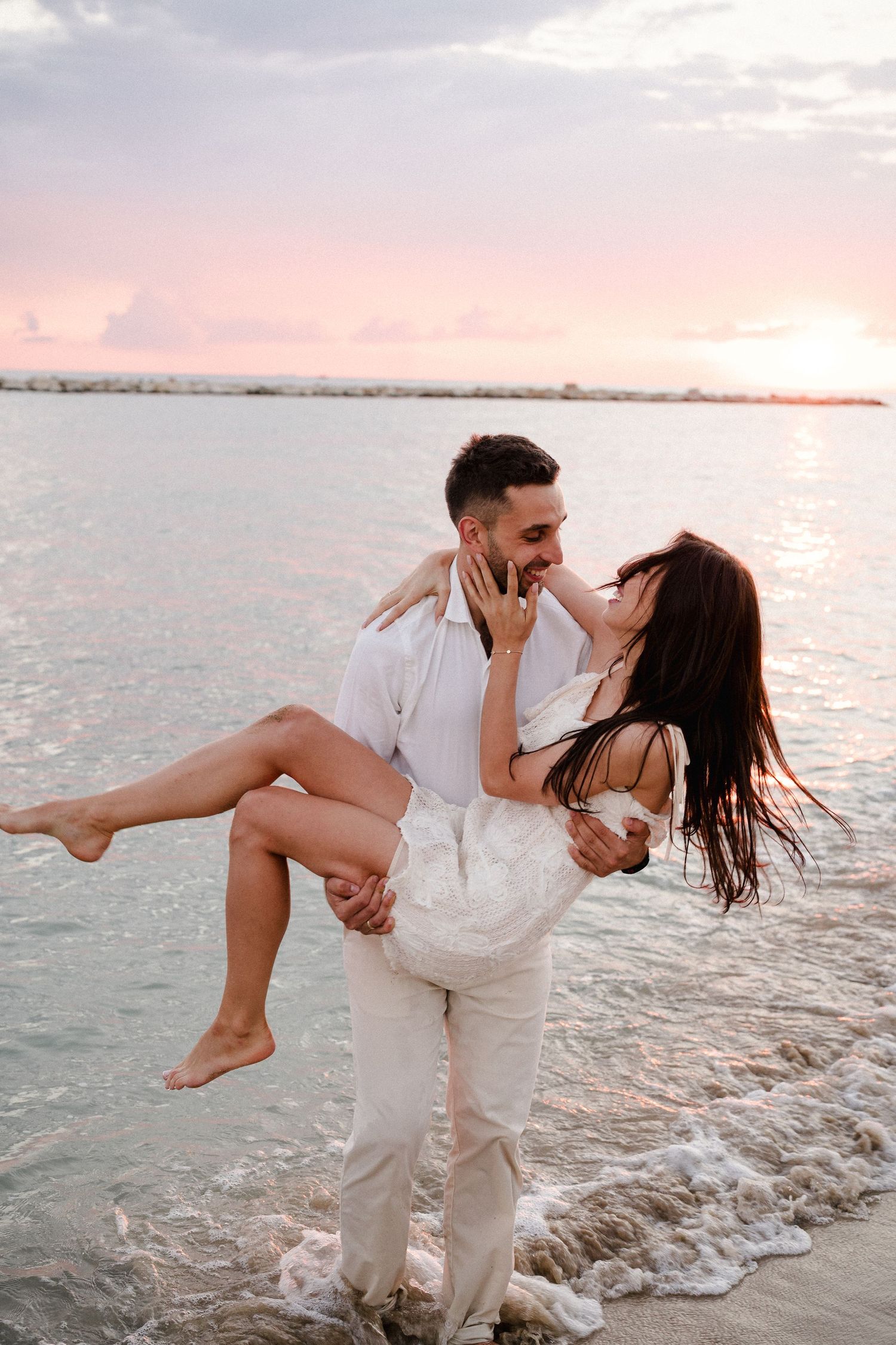 Romantic beach scene with couple in white embracing by ocean waves at sunset.
